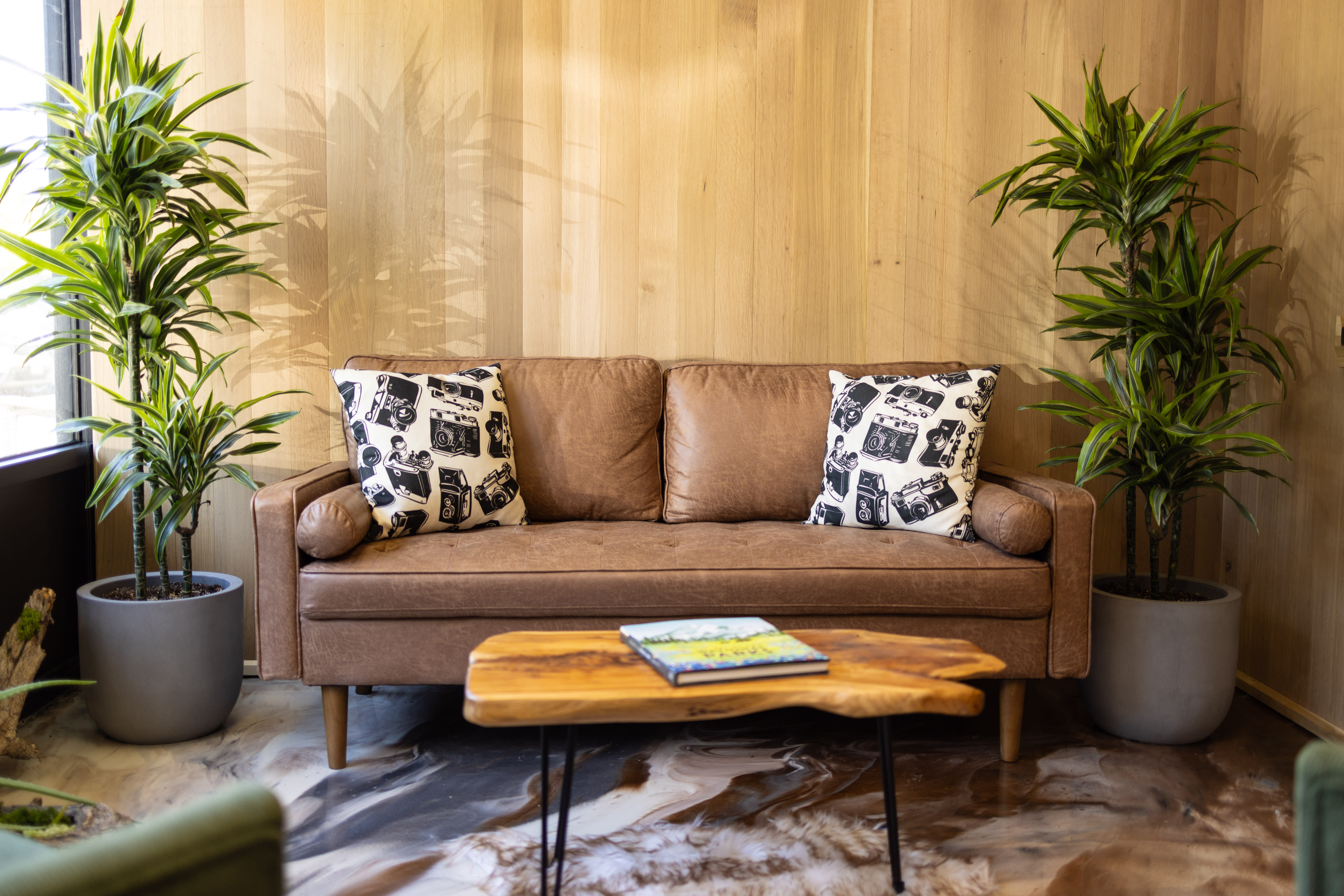 A brown sofa with patterned pillows sits in front of a wooden wall, flanked by potted plants and accompanied by a coffee table.