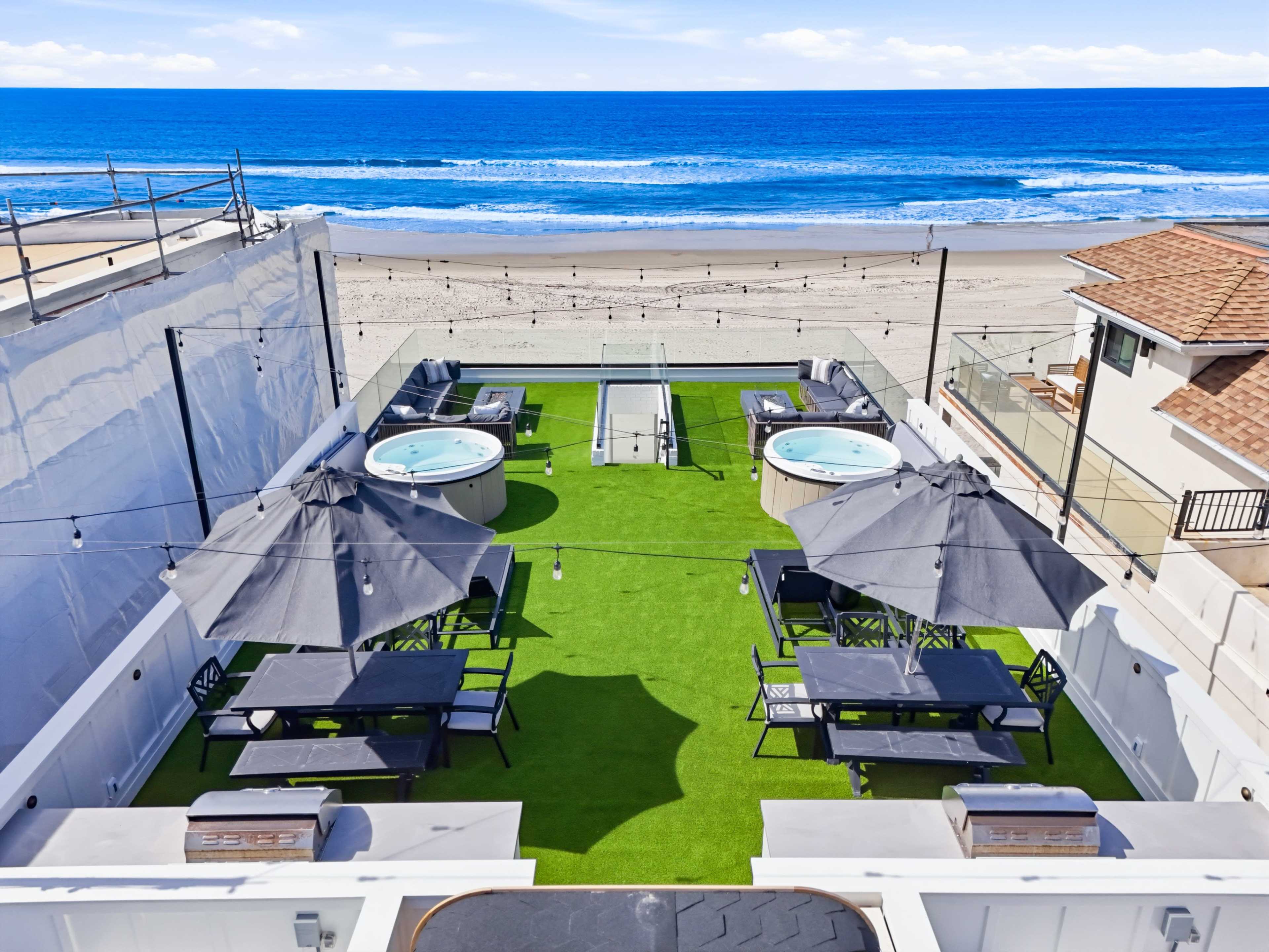 The image shows a rooftop terrace featuring several hot tubs, dining tables, and umbrellas, overlooking a beach and ocean.