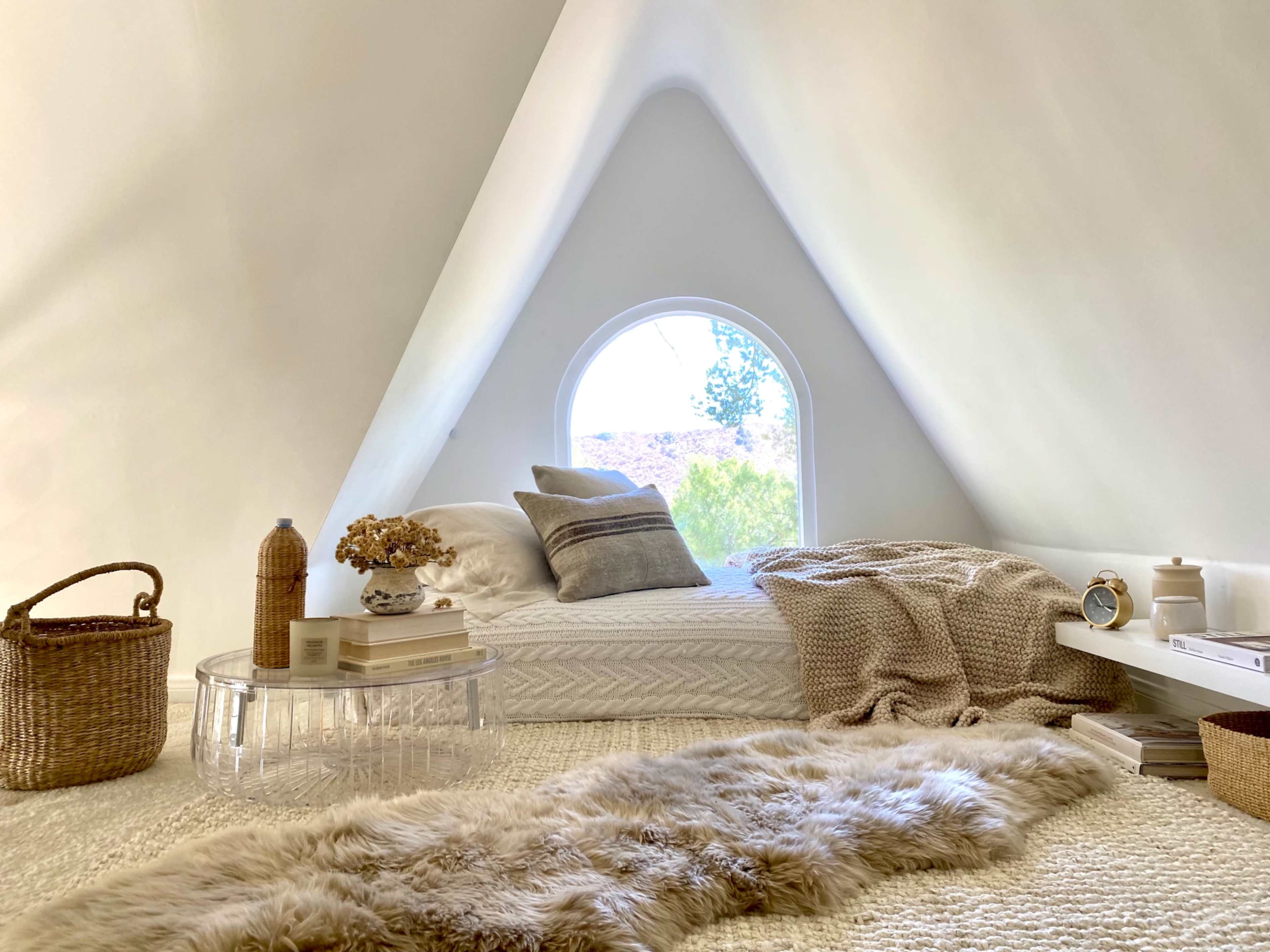 A minimalist bedroom with a sloped, white wall, a large arched window, and a bed adorned with neutral-colored blankets and decorative items.