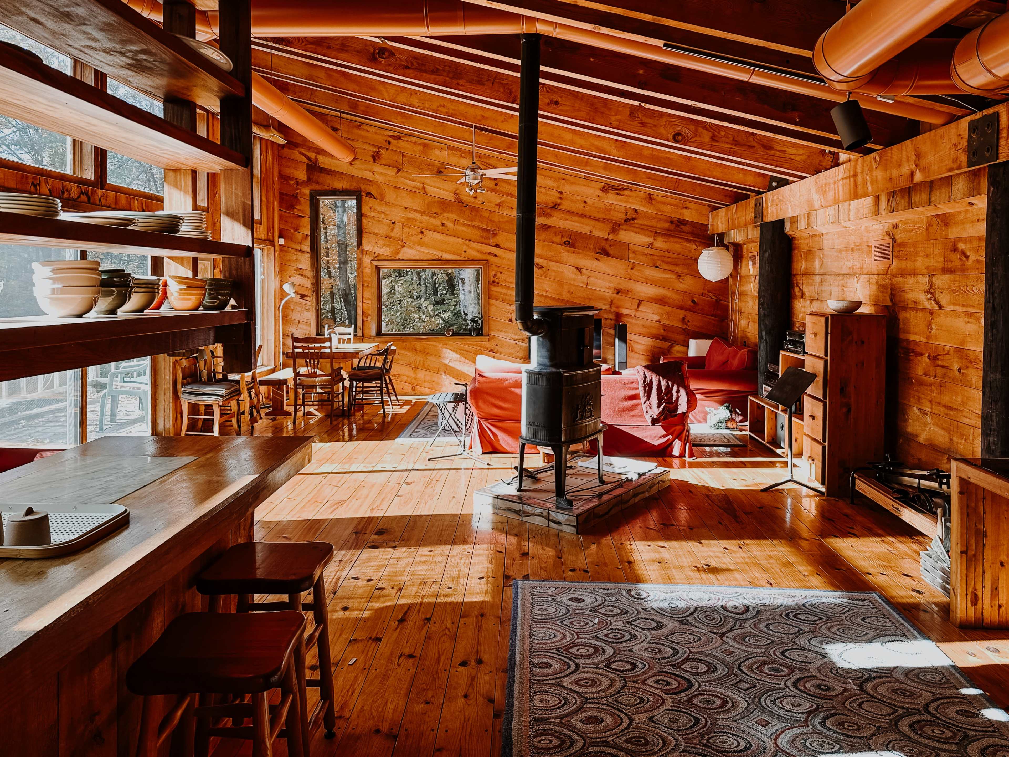 The image shows the interior of a wooden cabin with a cozy living area, featuring a central wood stove, a dining table, and large windows allowing natural light to enter.