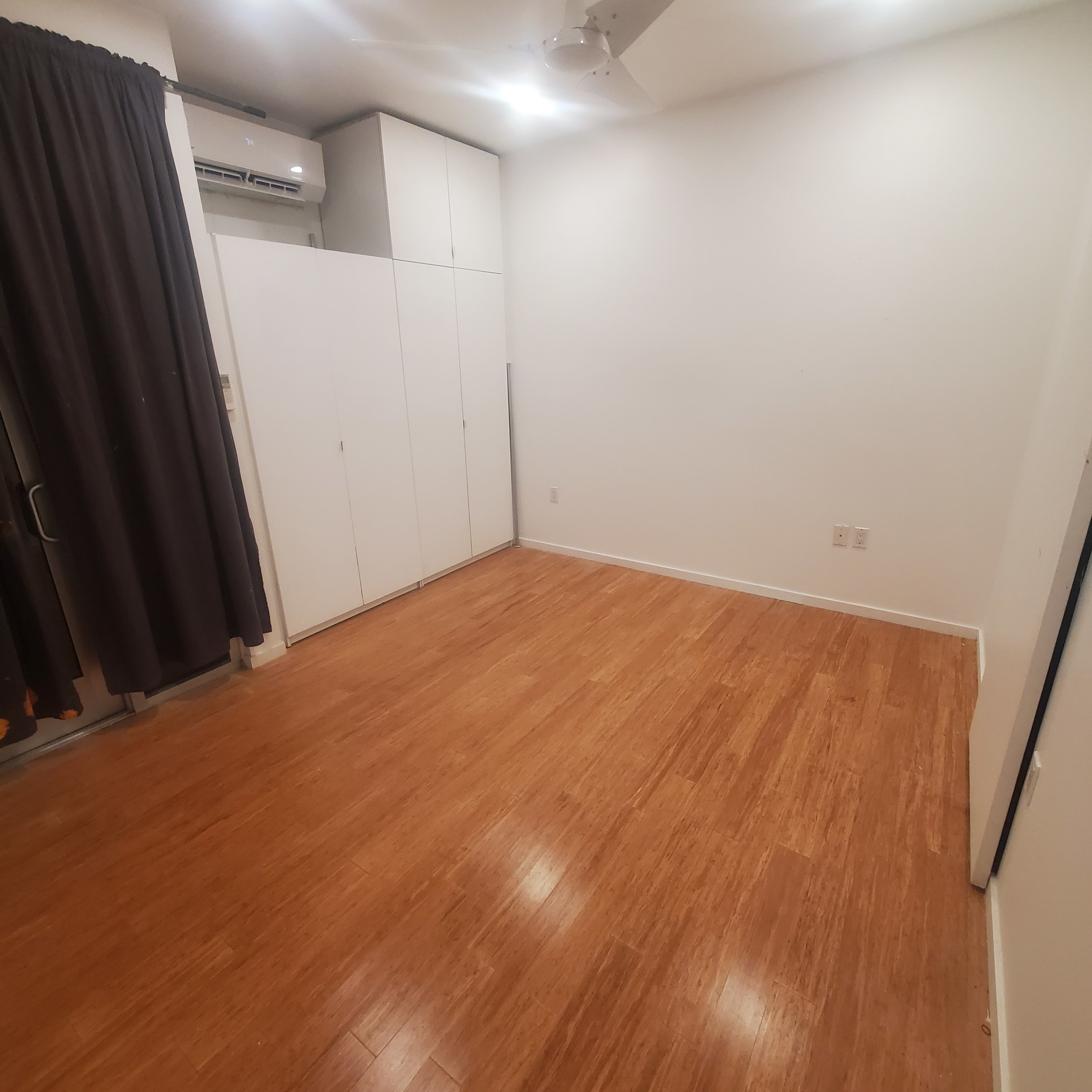 The image shows a vacant room with bamboo flooring, white walls, a ceiling fan, and a set of closet doors on one side.