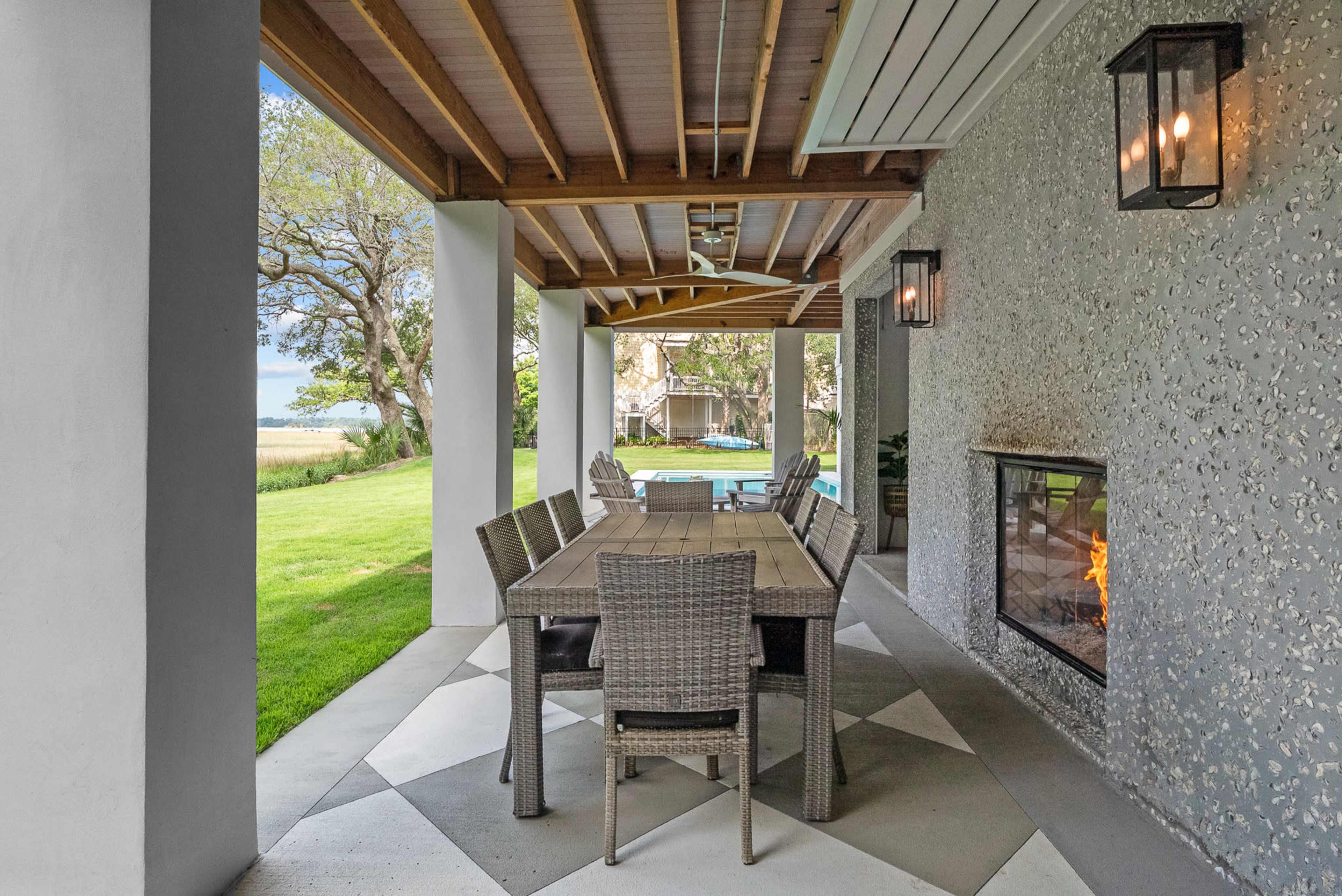 A spacious outdoor dining area features a long wooden table and wicker chairs under a covered patio, with a view of a green lawn and a pool in the background.