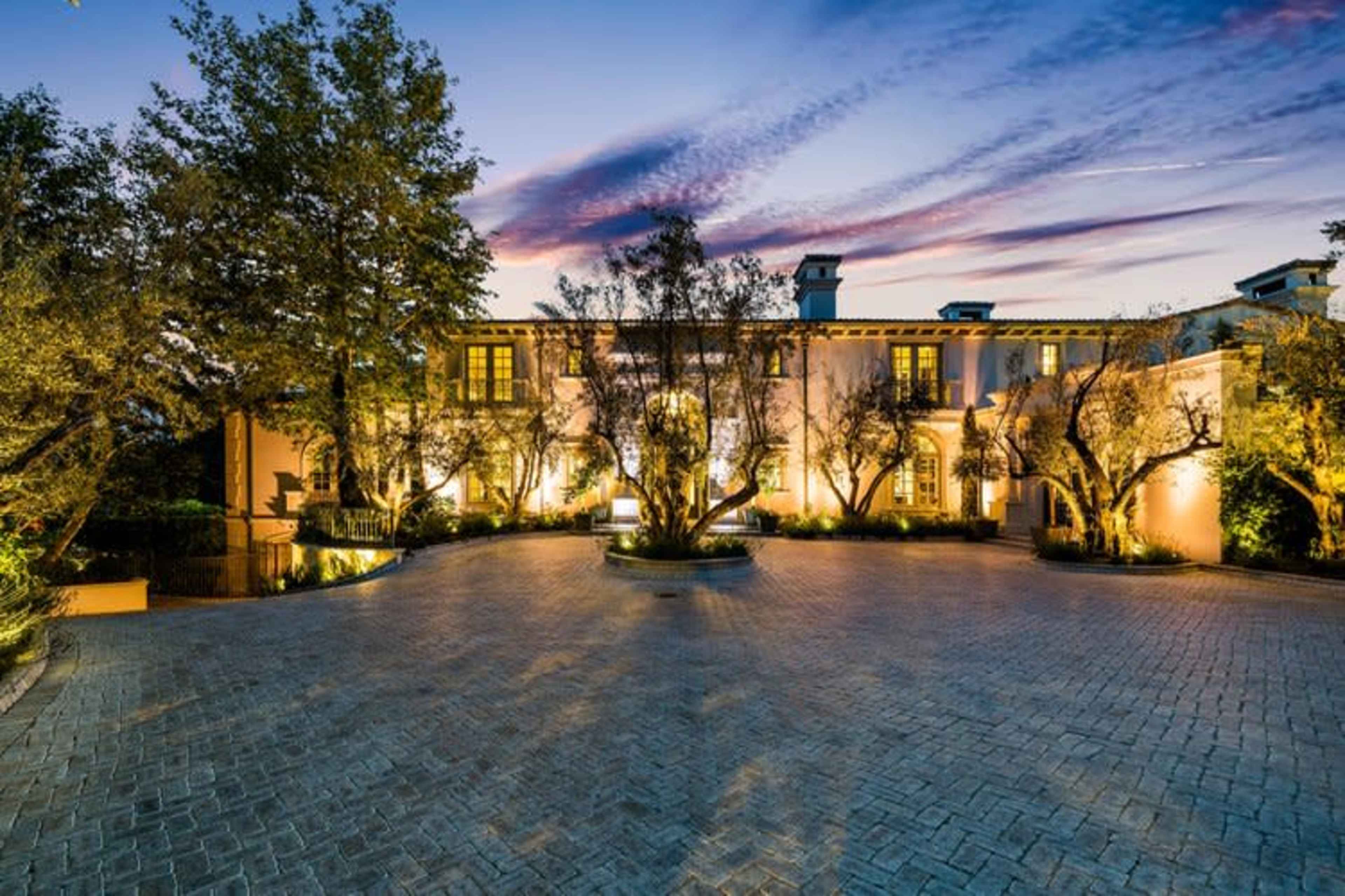 A large, elegantly designed mansion with a paved driveway is illuminated by exterior lights at twilight, surrounded by trees.