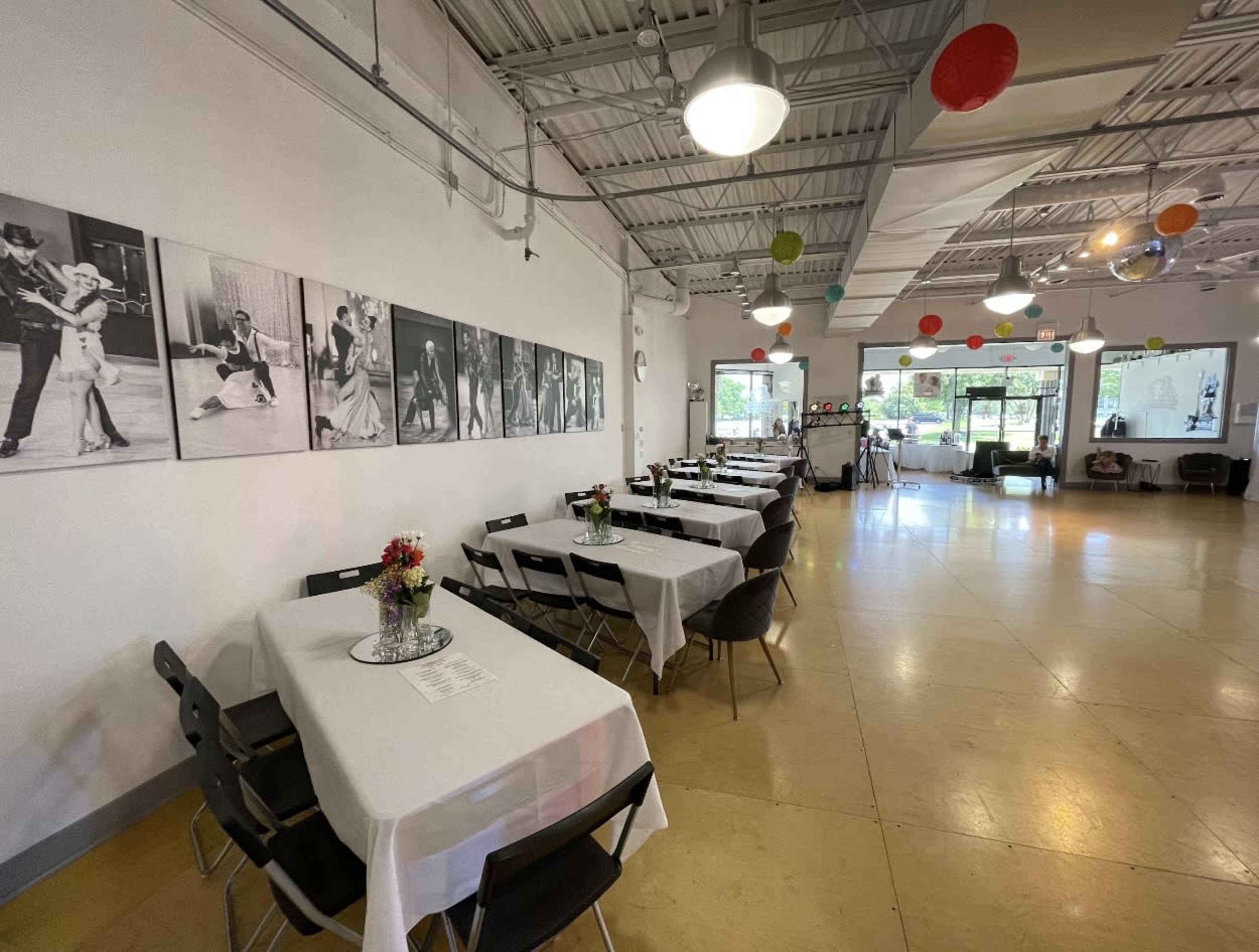 The image shows a spacious event venue with tables set for dining, black-and-white photographs of dancers on the wall, and colorful decorations hanging from the ceiling.