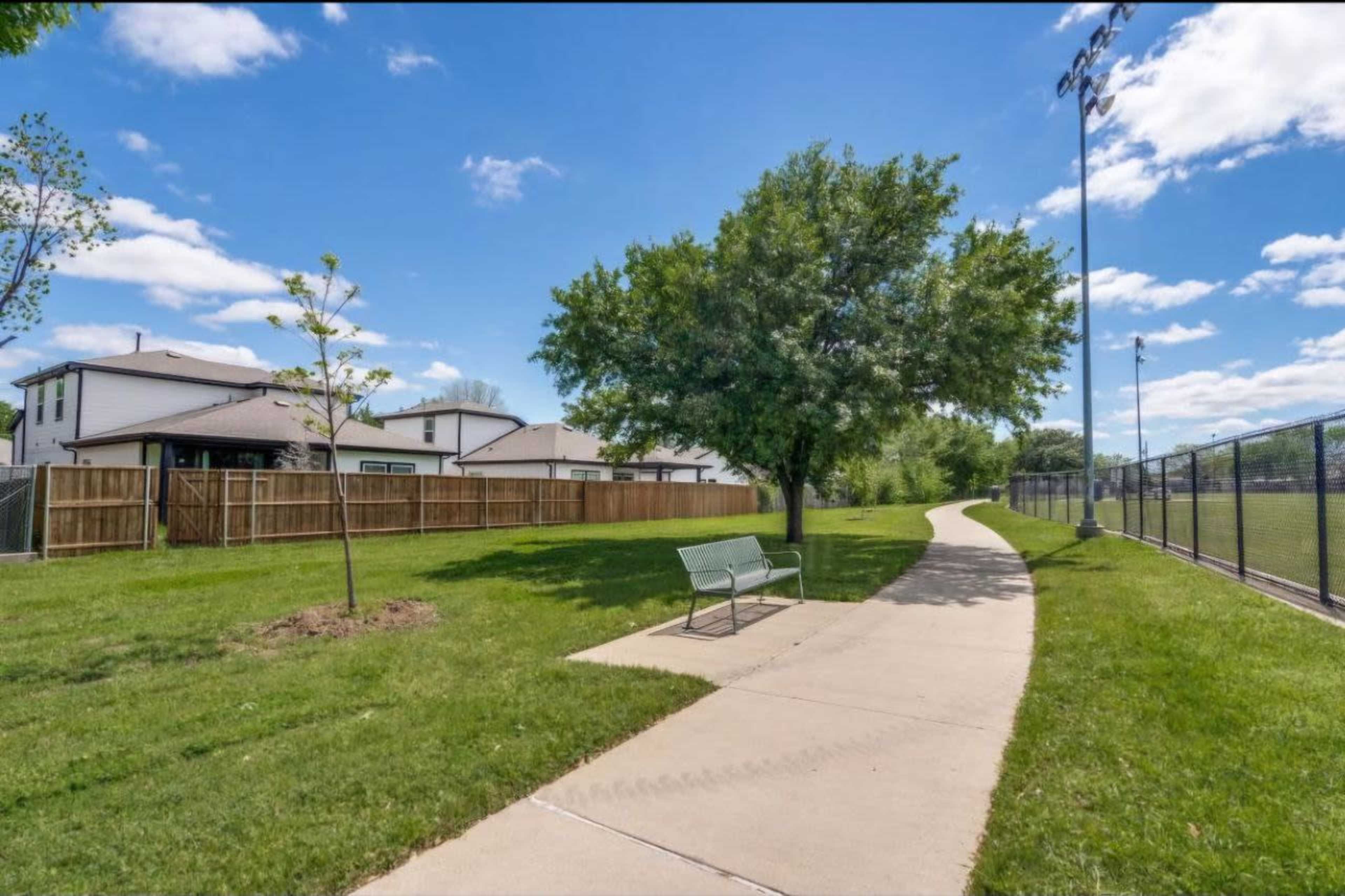 A concrete pathway curves alongside a grassy area and a fenced sports field, with a bench and a tree nearby.