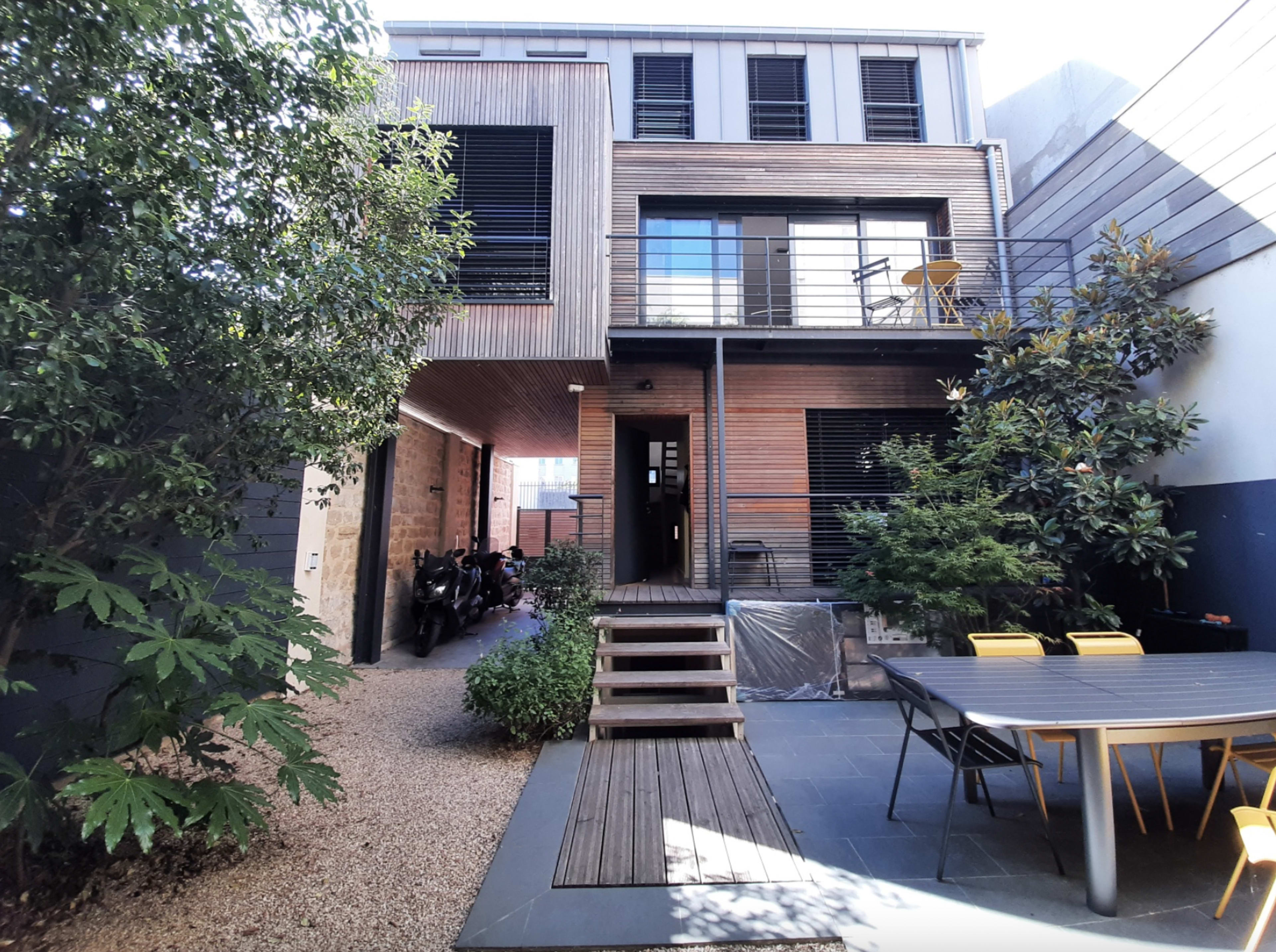 A modern house with a wooden exterior, a patio area, and a garden surrounded by plants and gravel pathways.
