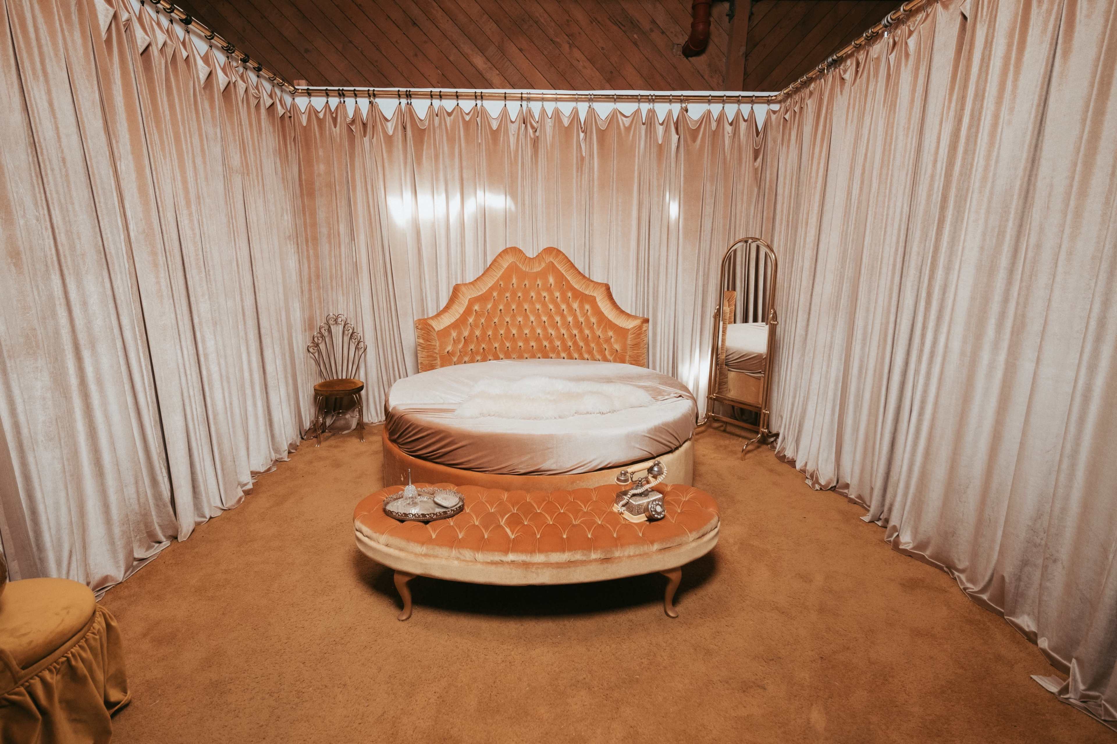 The image shows a vintage-style bedroom featuring a round, tufted orange bed surrounded by floor-length, cream-colored drapes and a small mirrored dressing table.