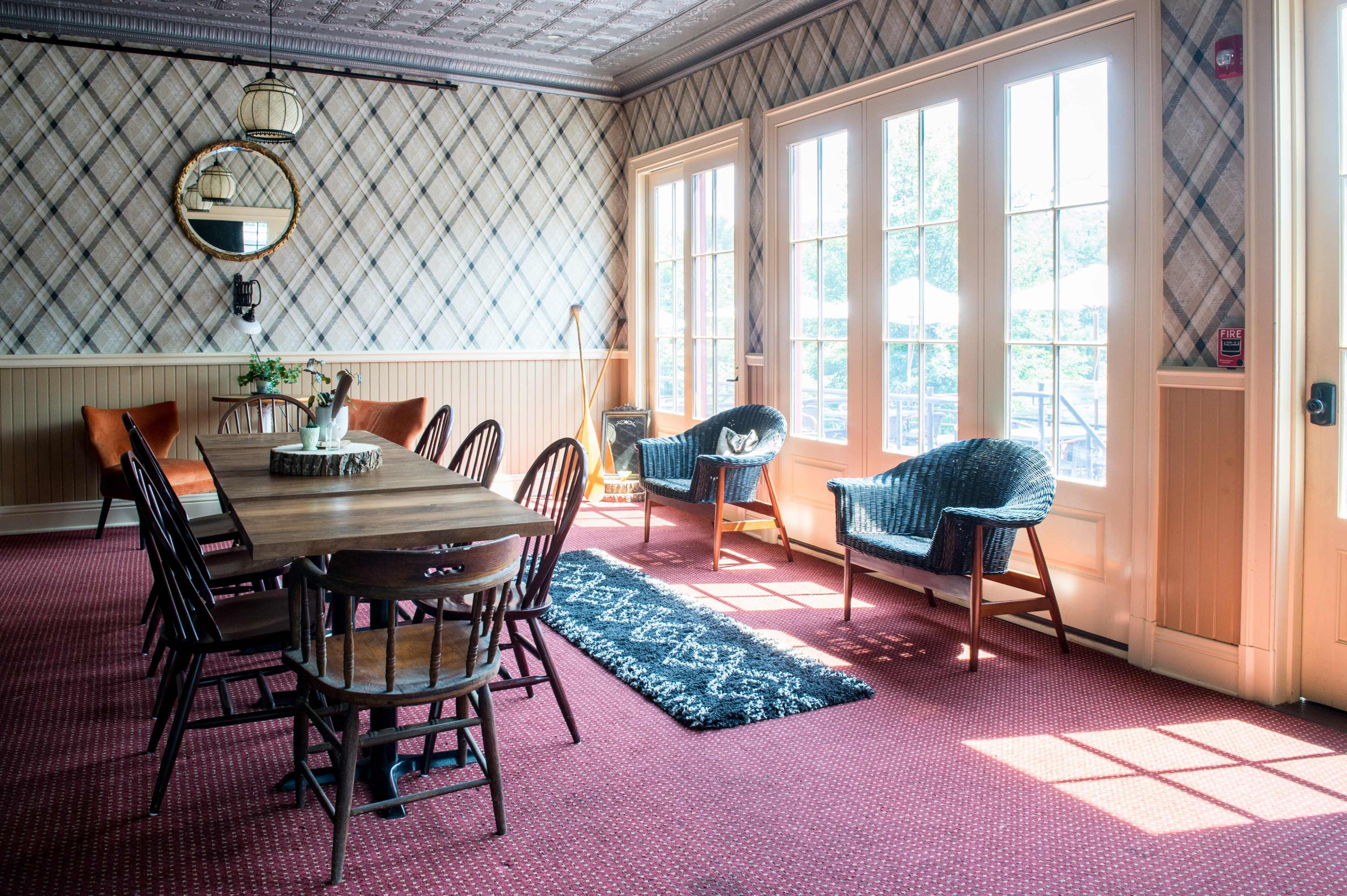 A well-lit room with a long wooden dining table surrounded by chairs, a patterned rug, and large windows that provide a view of an outdoor area.