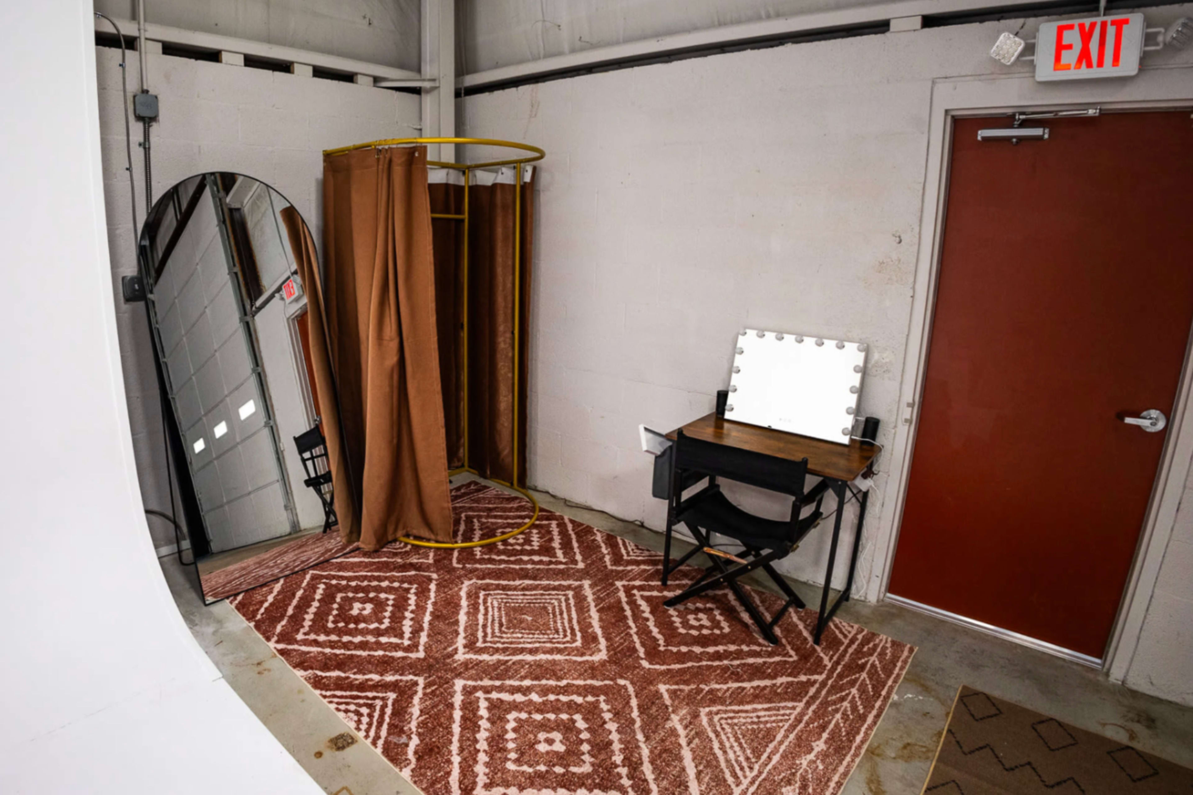 The image features a small room with a mirror, a curtain setup, a vanity table with lights, and an exit door.