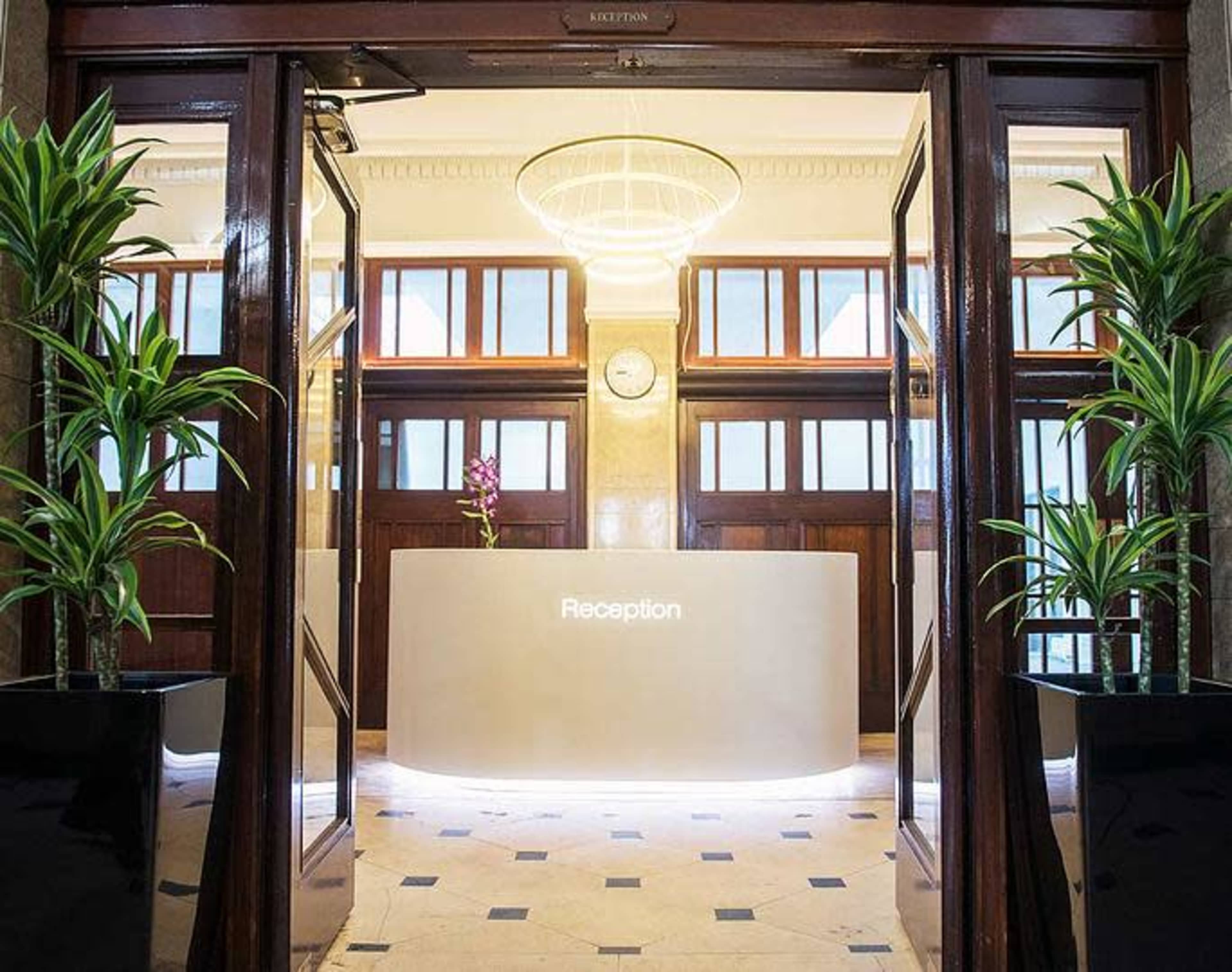 The image shows a modern reception area with a white reception desk, potted plants, and large glass doors leading into the space.