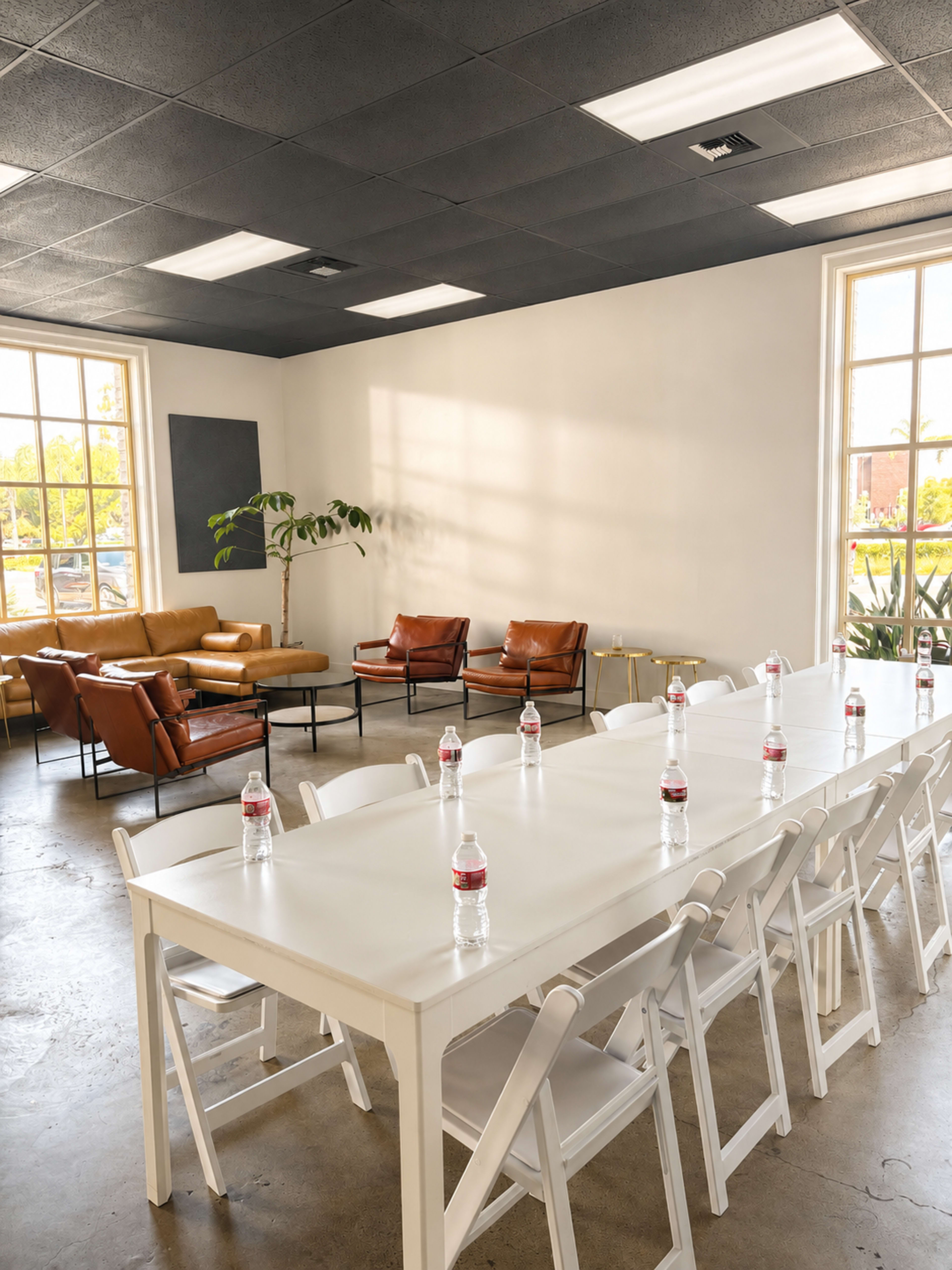The image shows a modern meeting room with a long white table set for a gathering, surrounded by chairs and featuring a seating area with brown leather sofas and a potted plant.