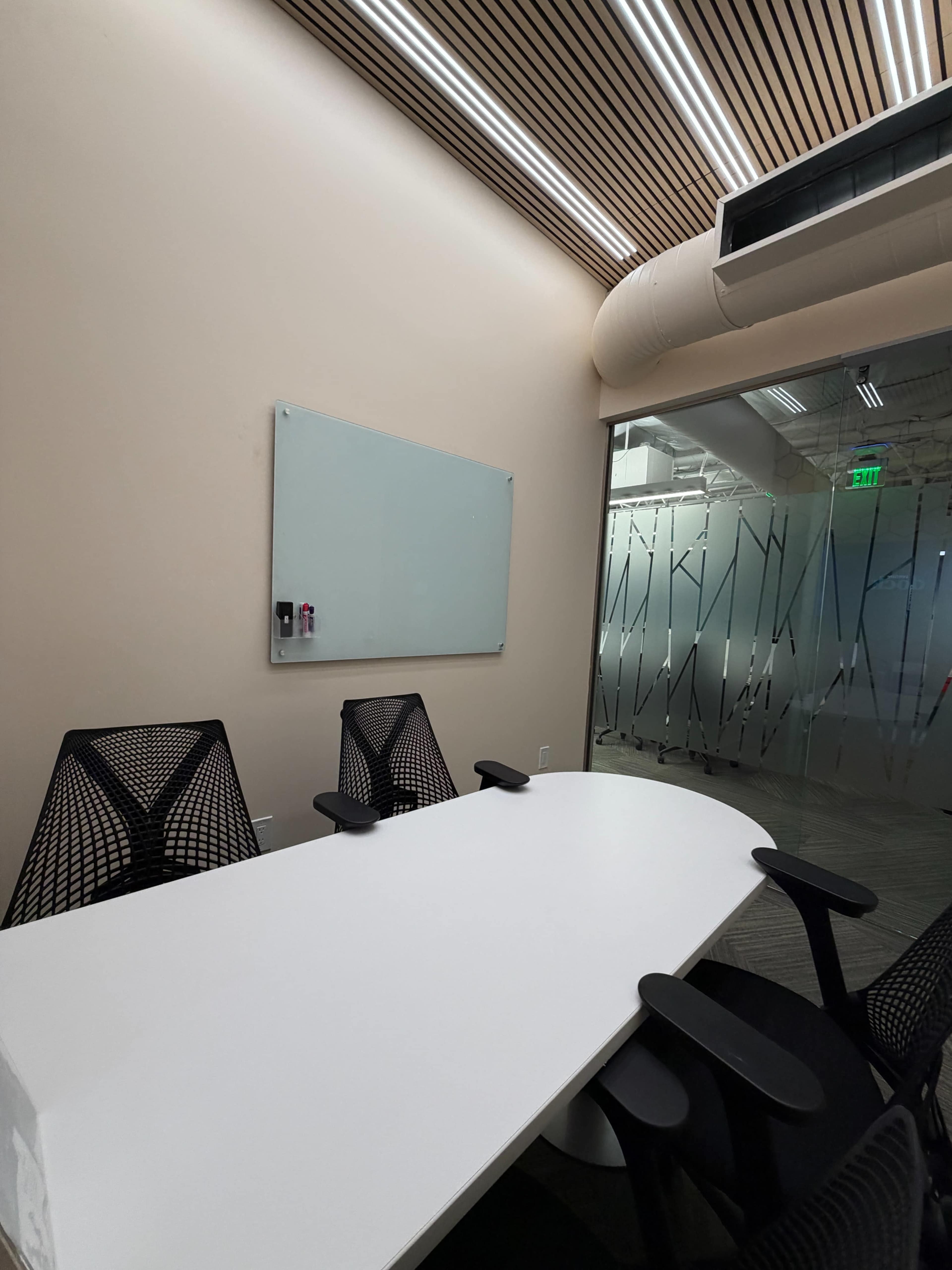 A modern conference room features a white oval table surrounded by ergonomic chairs, with a whiteboard on the wall and glass doors in the background.