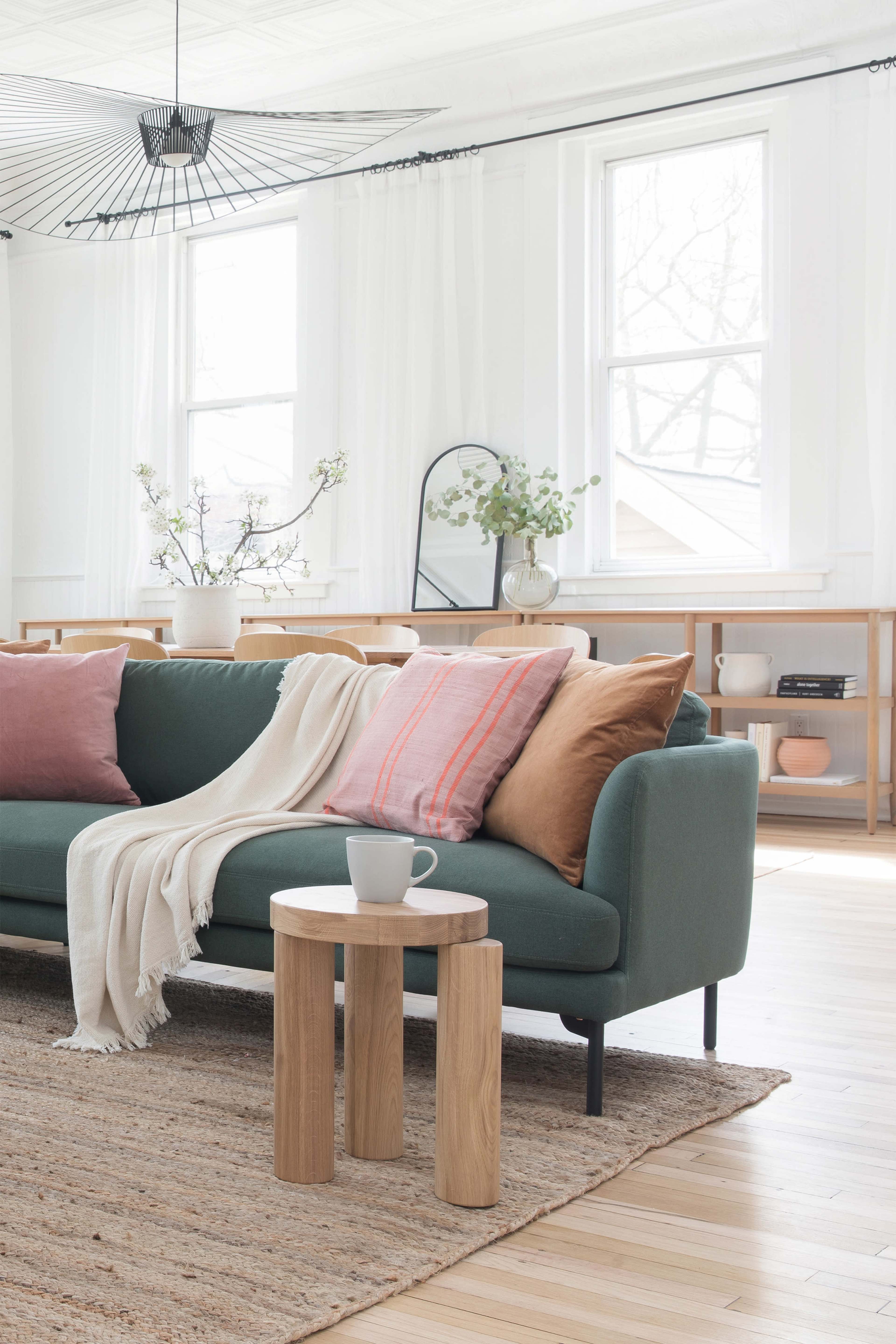 A green sofa with decorative pillows sits beside a small wooden side table in a well-lit, modern living room.