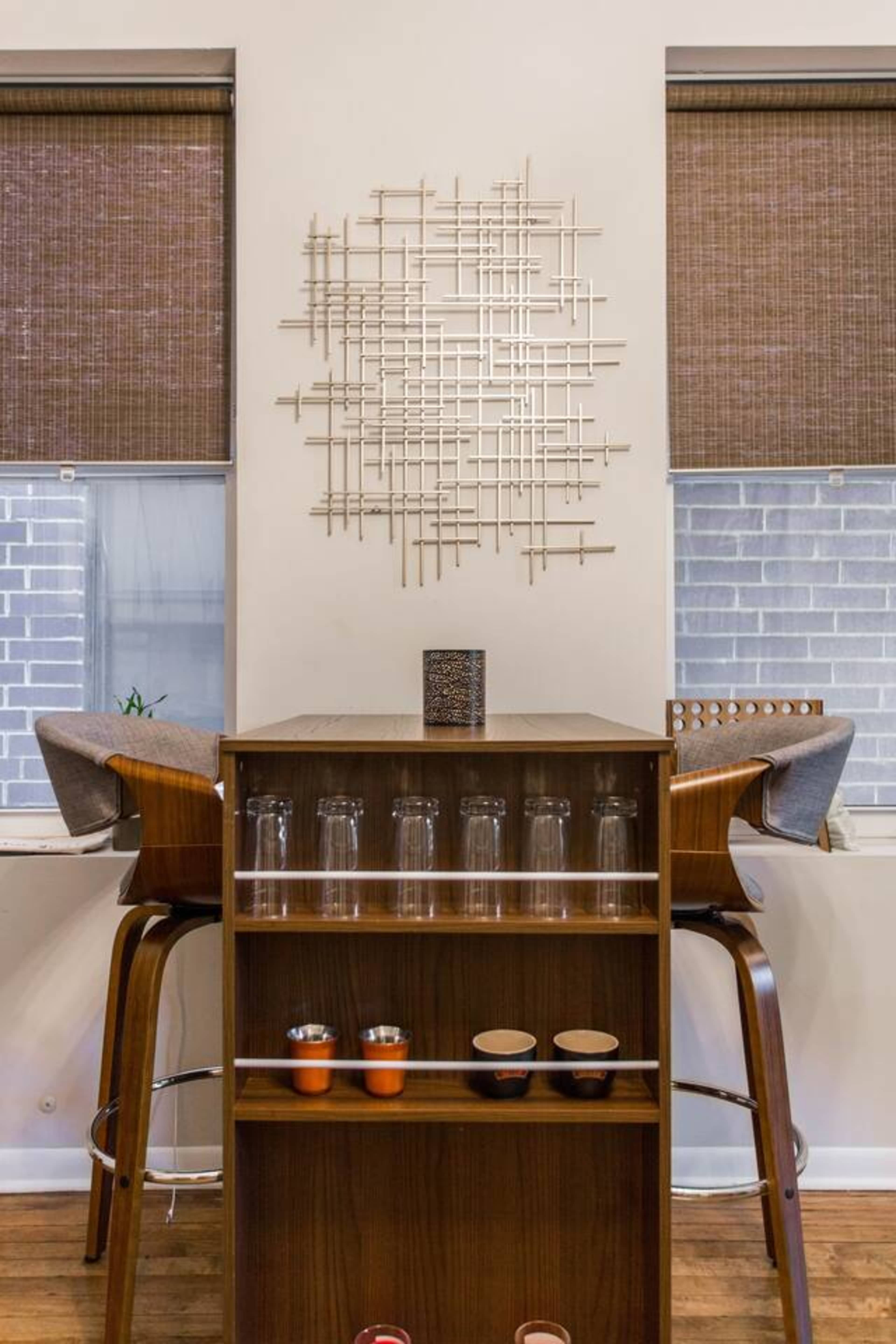 A modern kitchen area featuring a wooden bar unit with glassware and decorative items, paired with two high-backed stools and textured window shades.