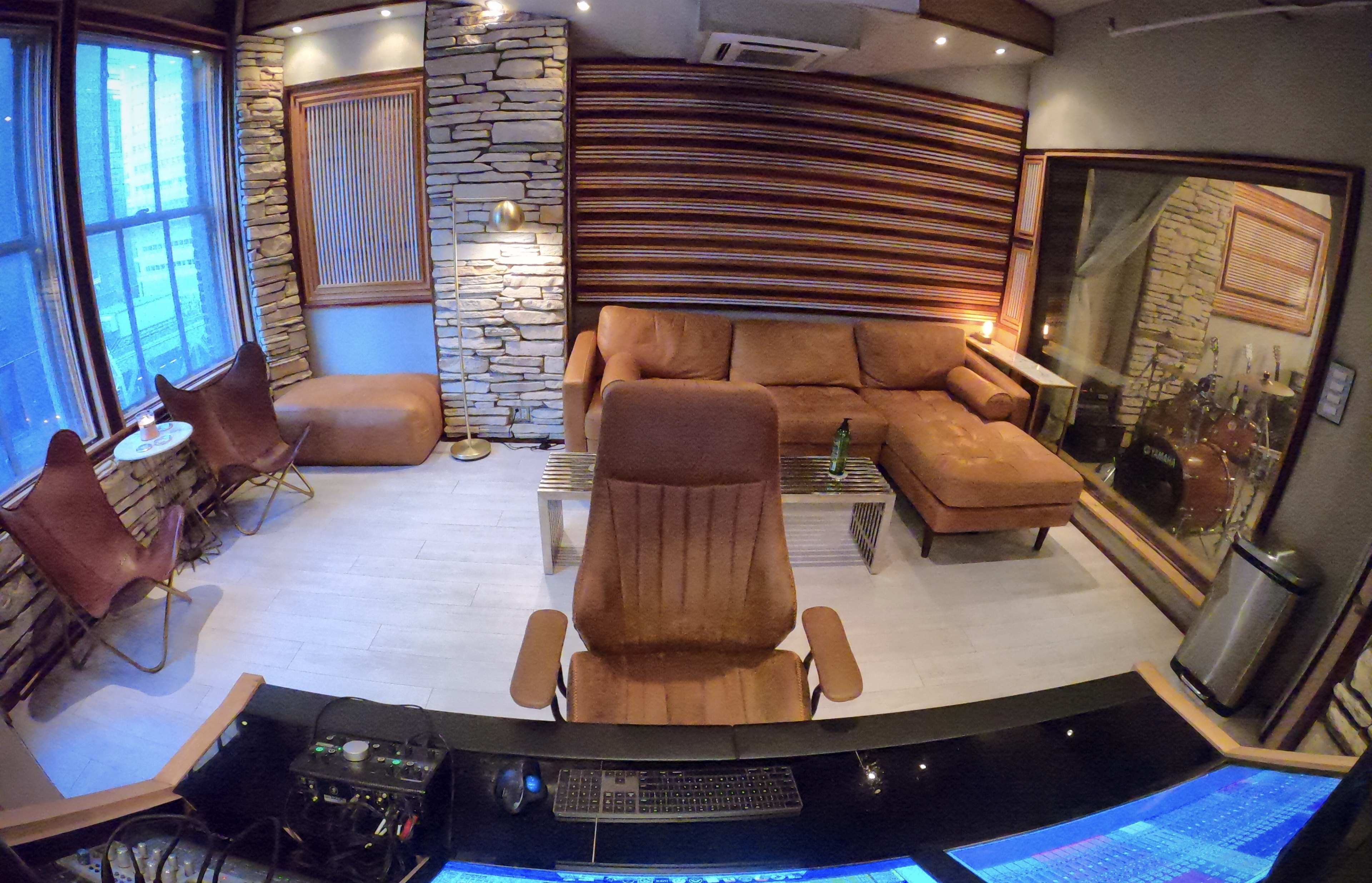 A cozy room with stone walls, a brown sectional sofa, a recording studio setup, and large windows allowing natural light.