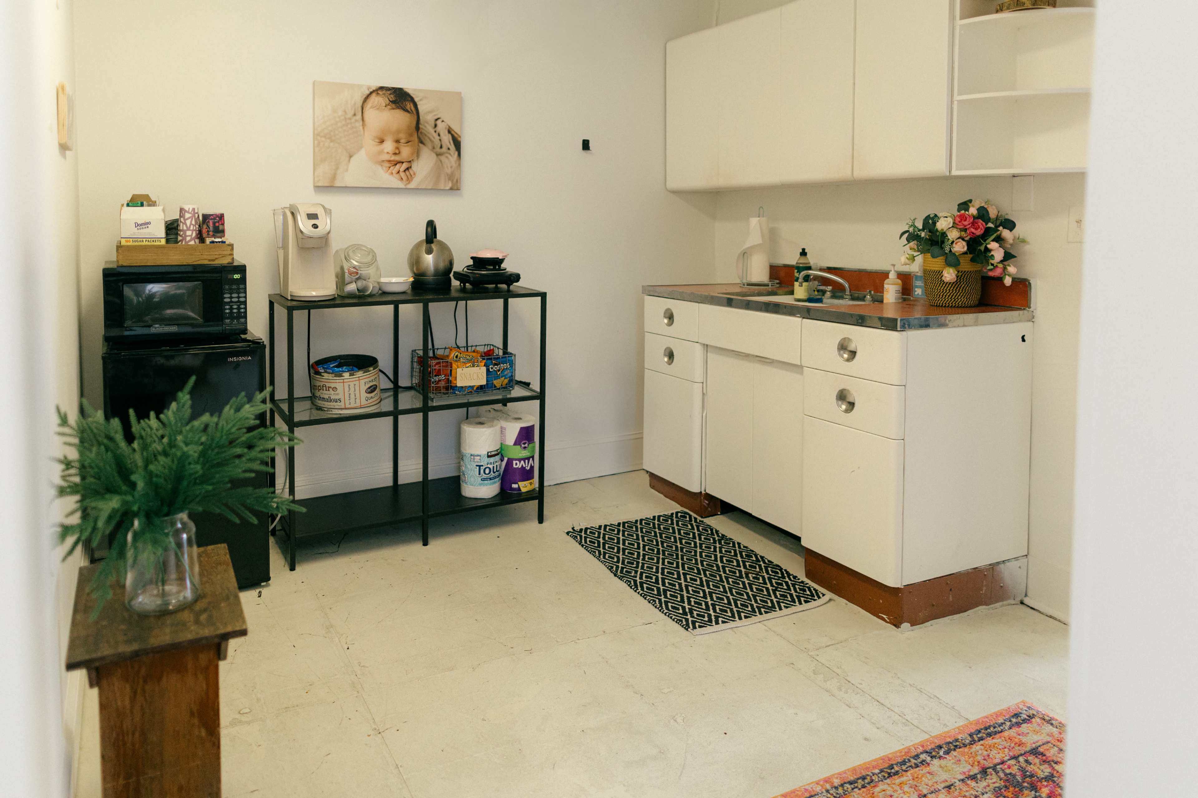 The image shows a small kitchen with minimal cabinetry, a black metal shelf holding kitchen items, and a sink against a light-colored wall.