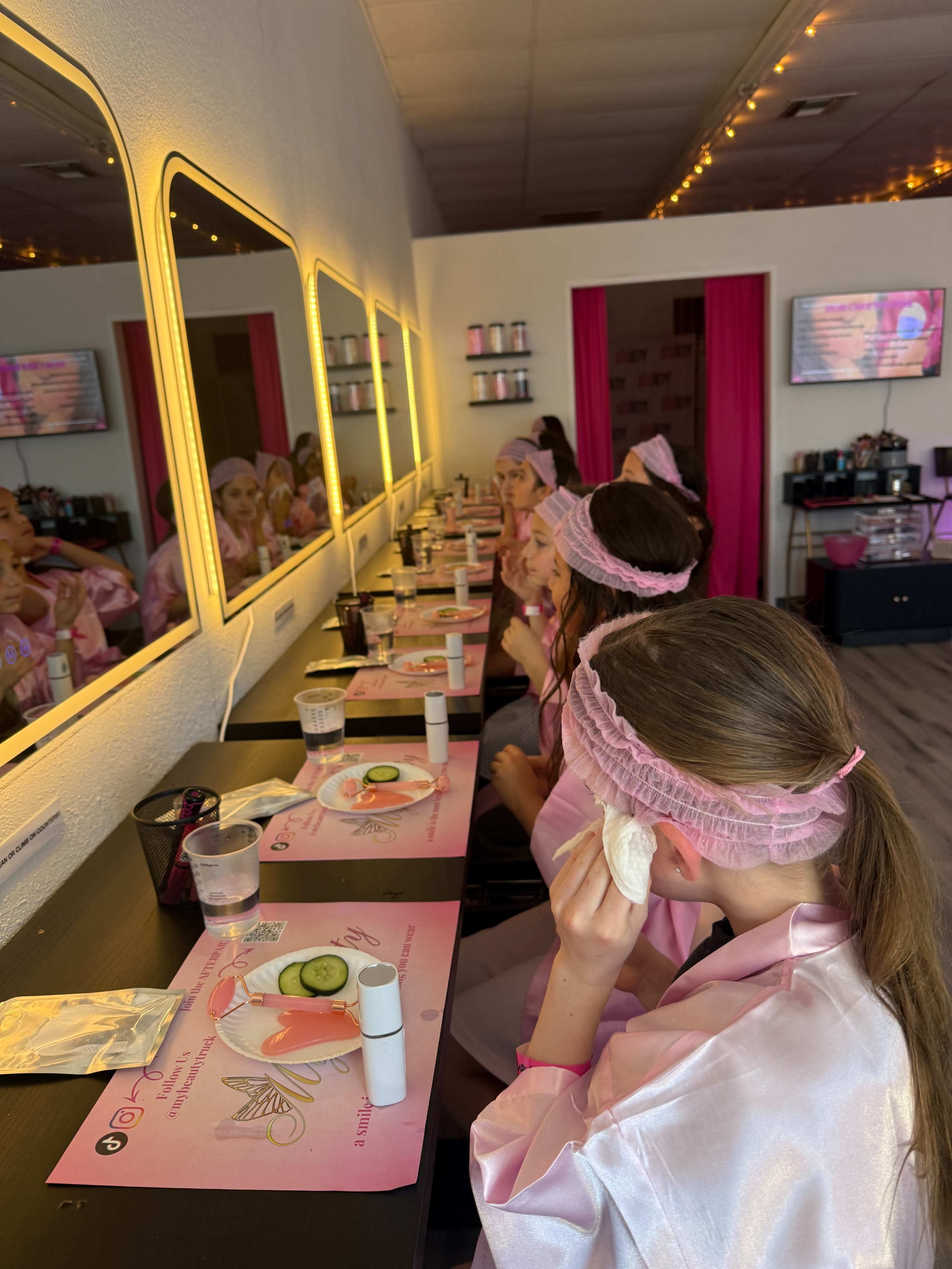 A group of girls in matching pink robes and headbands are sitting at a vanity with mirrors, preparing for a spa treatment that includes applying cucumber slices to their faces.