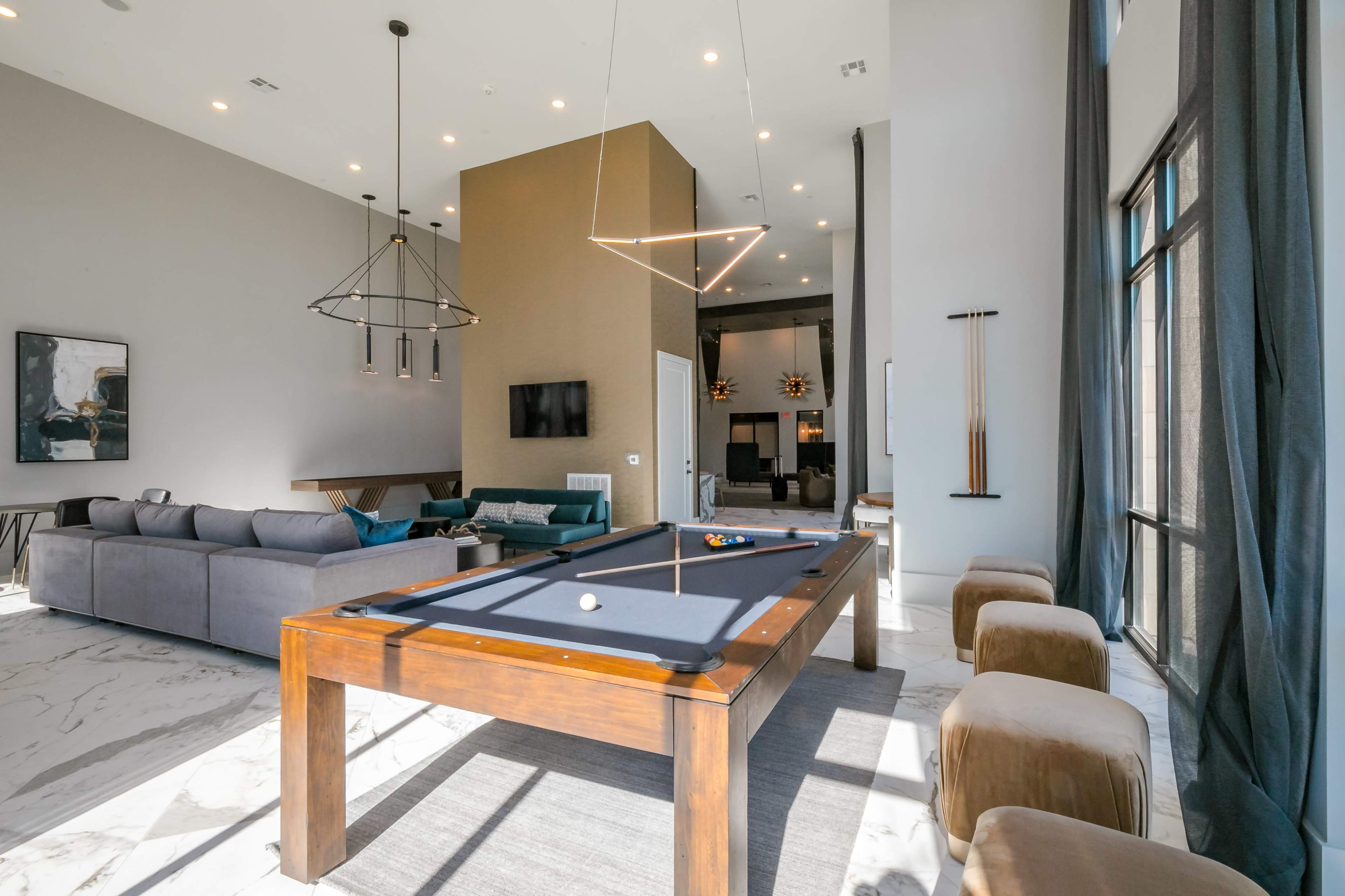 The image shows a modern lounge area featuring a pool table, a large sectional sofa, and tall windows that allow natural light to fill the space.
