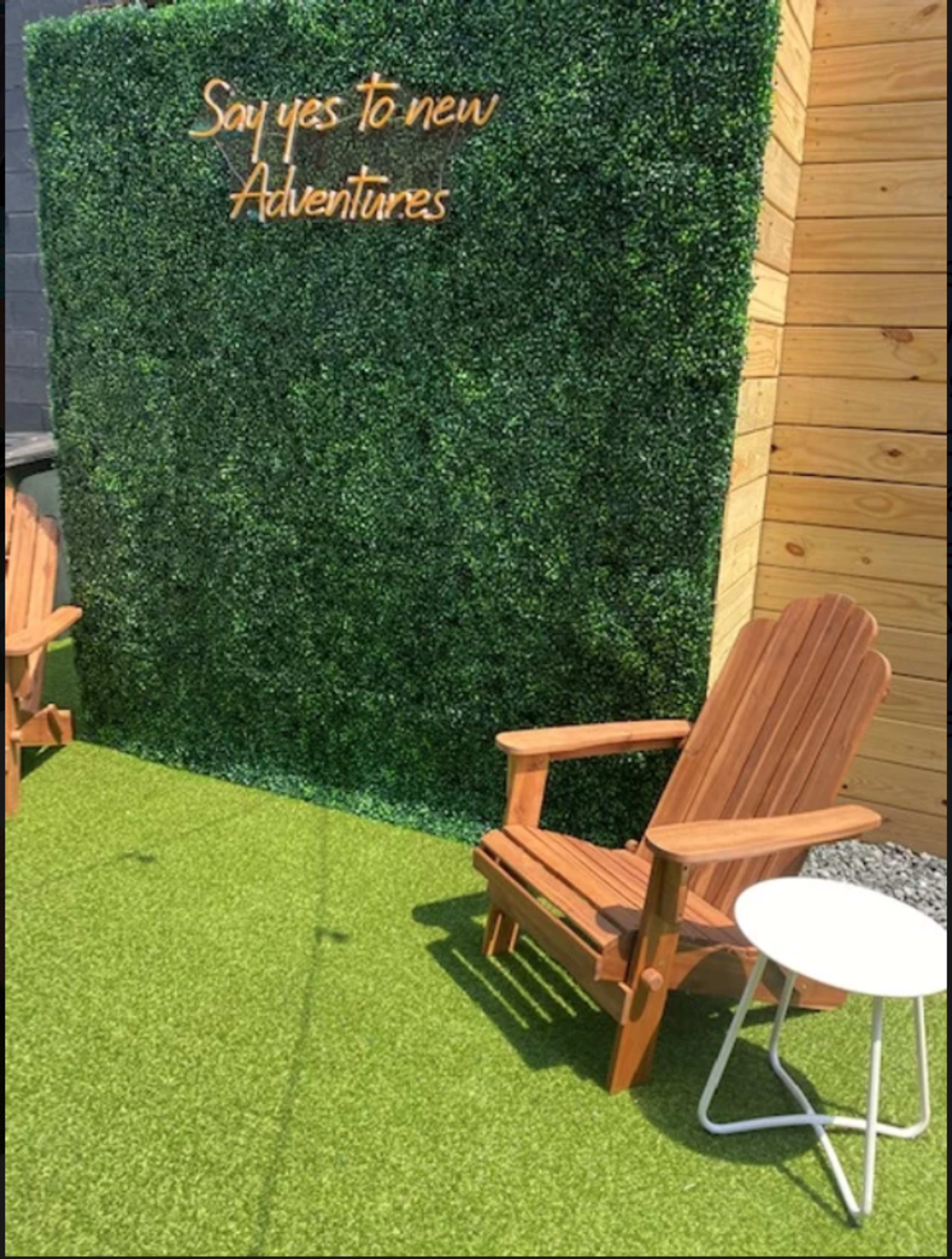 Two wooden chairs and a small white table in front of a green artificial hedge with the phrase "Say yes to new adventures" displayed above.
