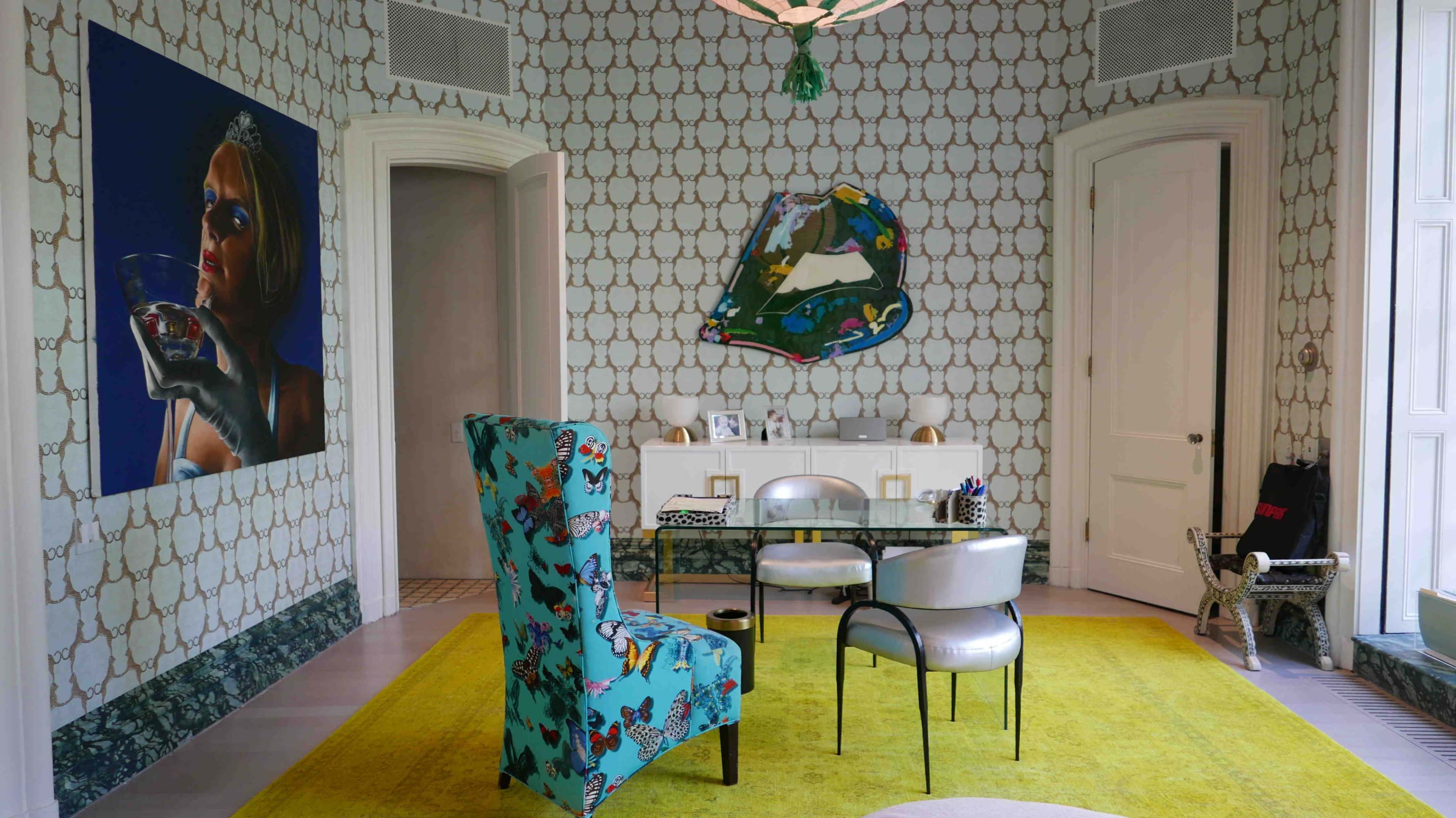 The image shows a stylish room with patterned wallpaper, a colorful chair, a white rug, a decorative table, and a large framed artwork on the wall.