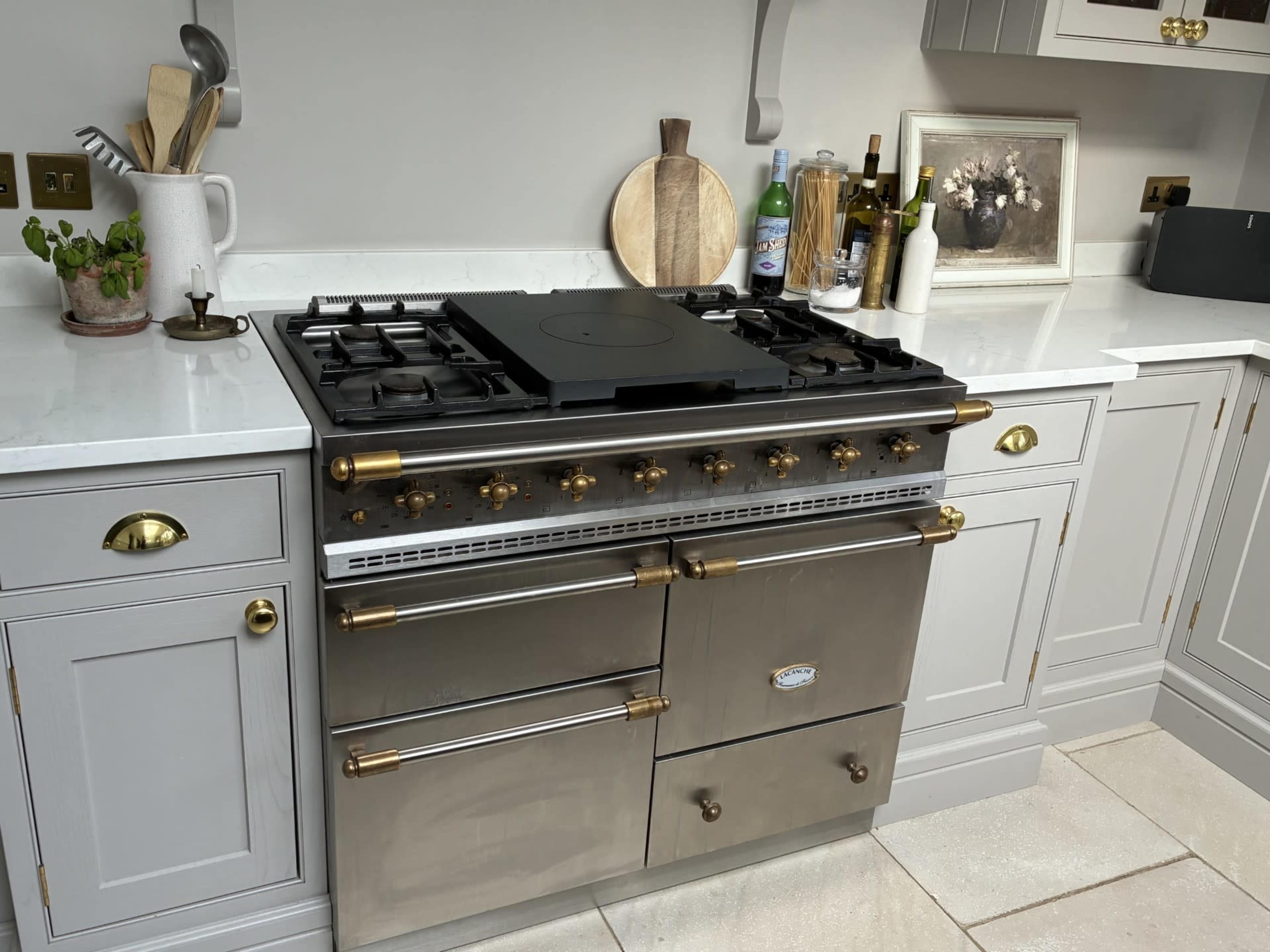 The image shows a stainless steel kitchen range with multiple burners, situated in a modern kitchen with gray cabinetry and a marble countertop.