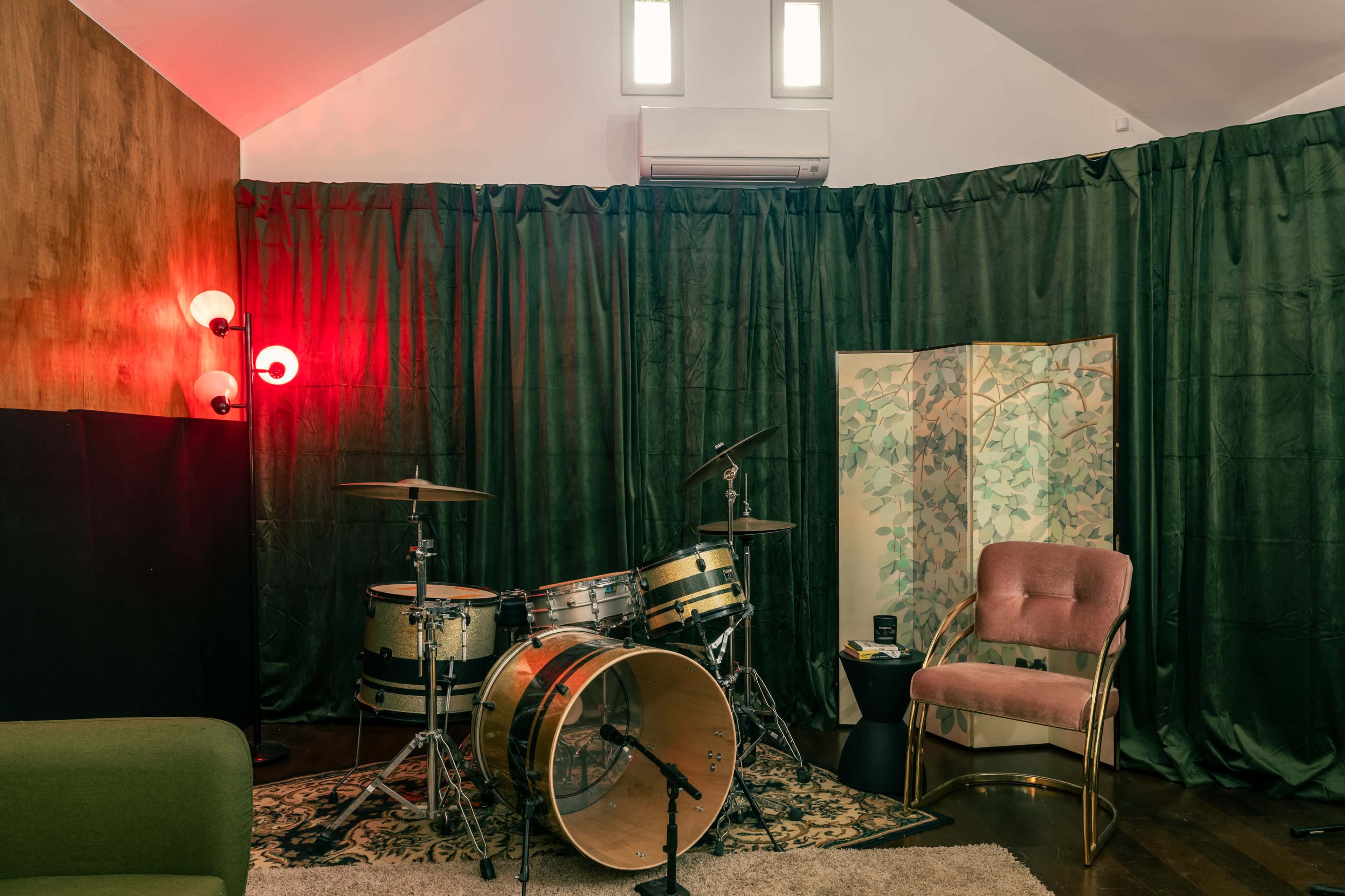 The image shows a small music studio featuring a vintage drum set, a pink armchair, and green curtains, with a red accent light illuminating the space.