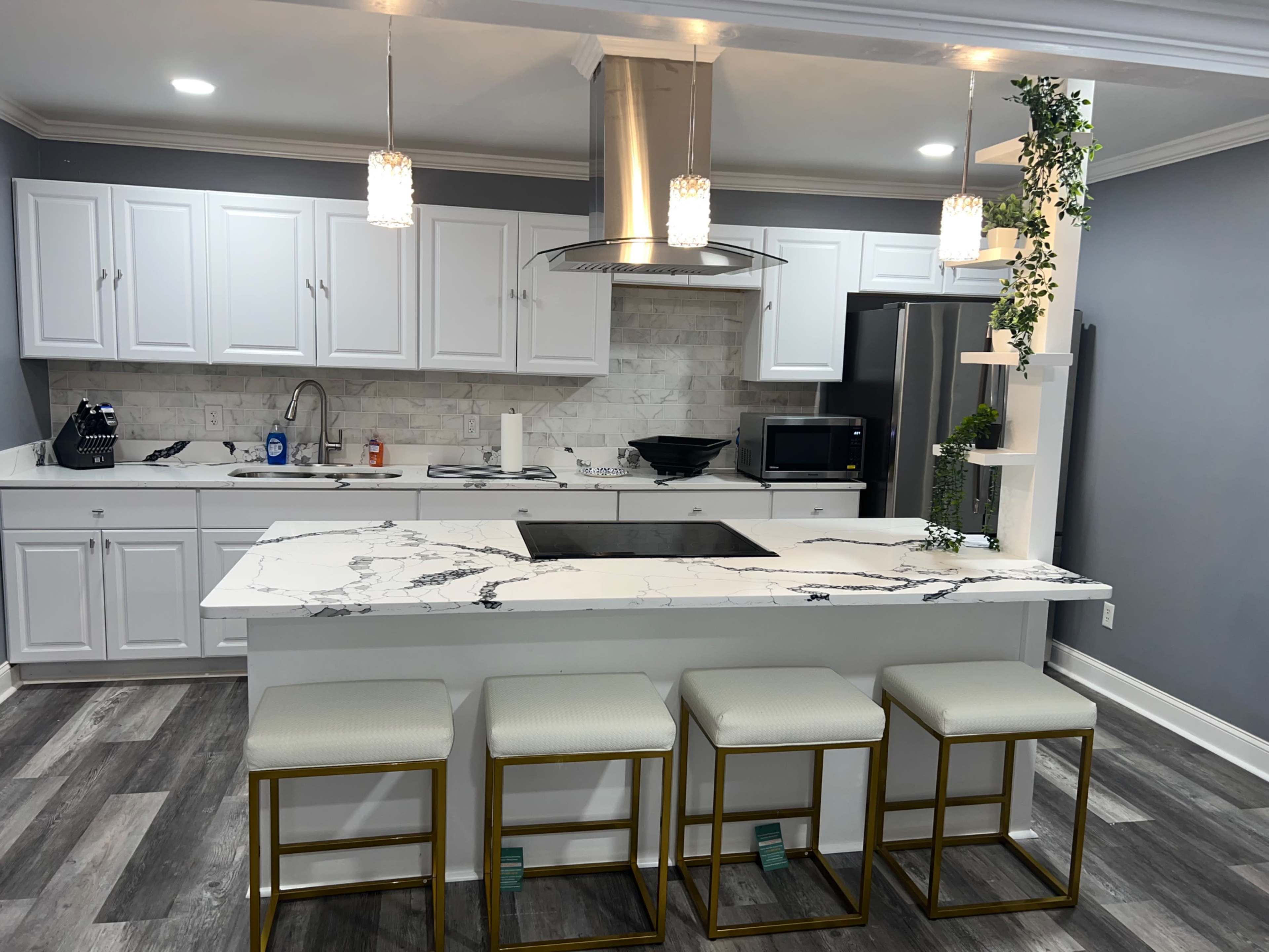 A modern kitchen features white cabinetry, a marble countertop with an integrated cooktop, and four gold-framed stools at the island.