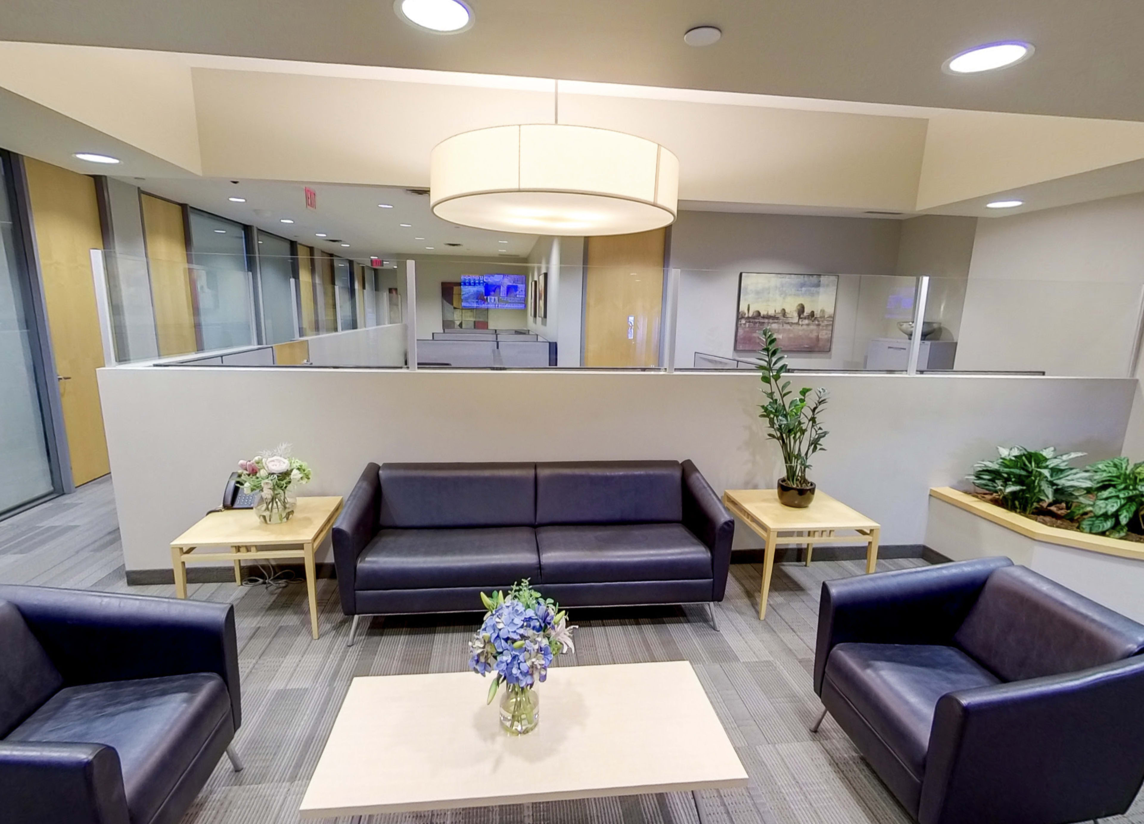 A modern waiting area features leather sofas and wooden tables, with plants and floral arrangements decorating the space.