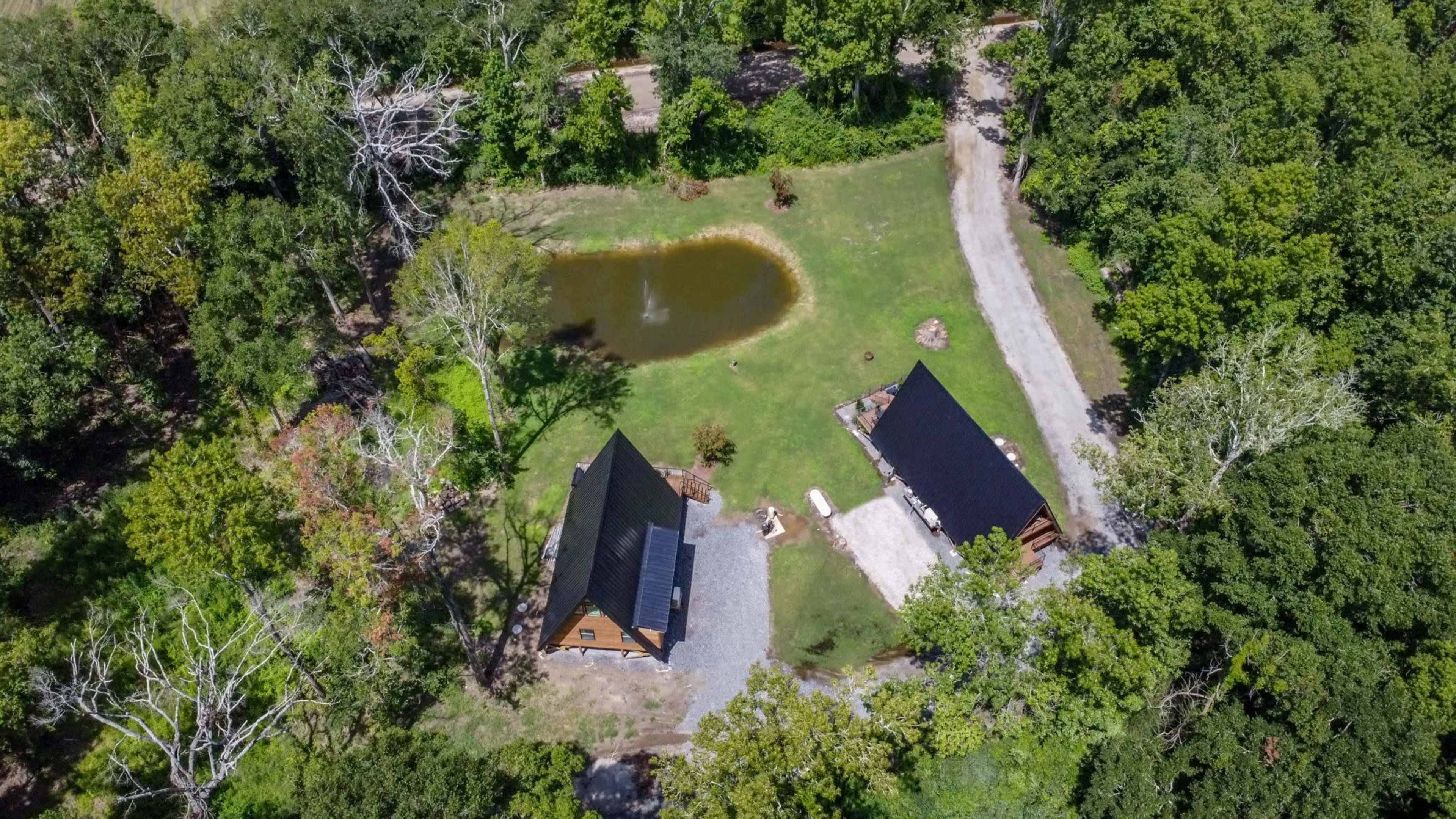 An aerial view shows a wooded area with a small pond and two cabins near a gravel pathway.