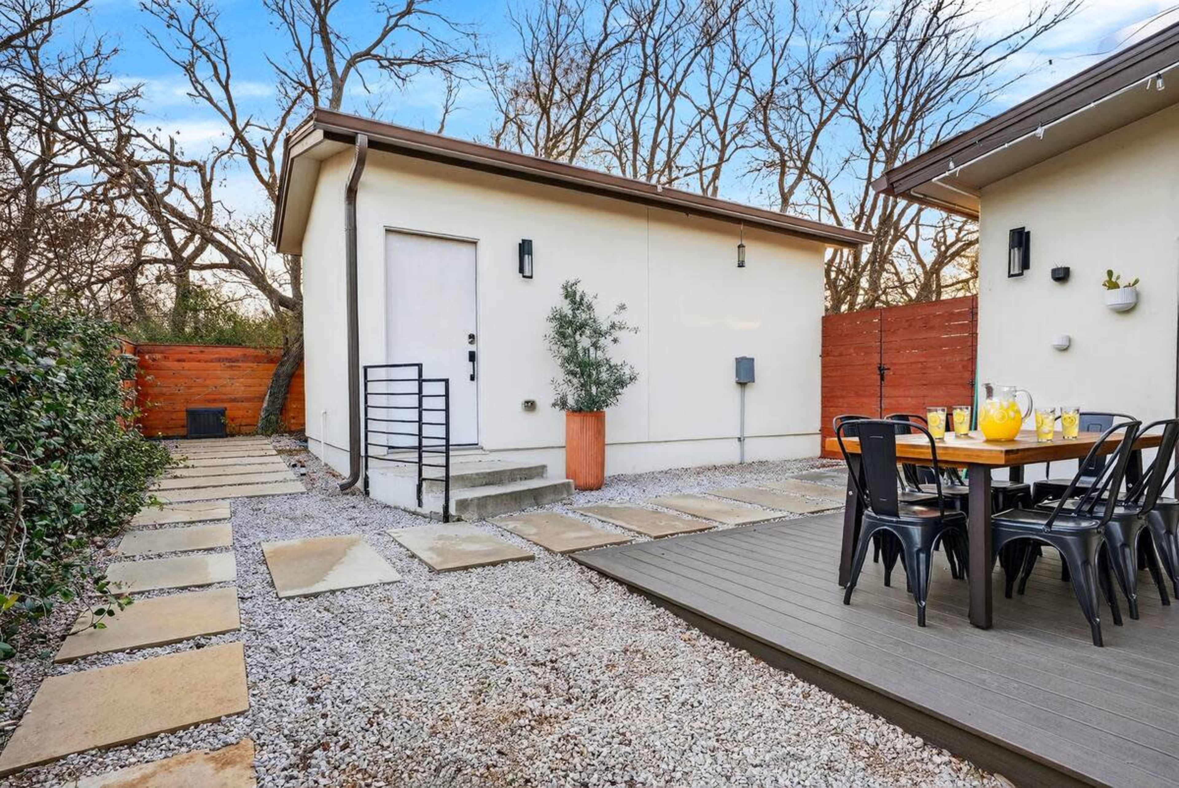 The image shows a stone pathway leading to a white building with a door, surrounded by gravel, with a table and chairs set for dining nearby.