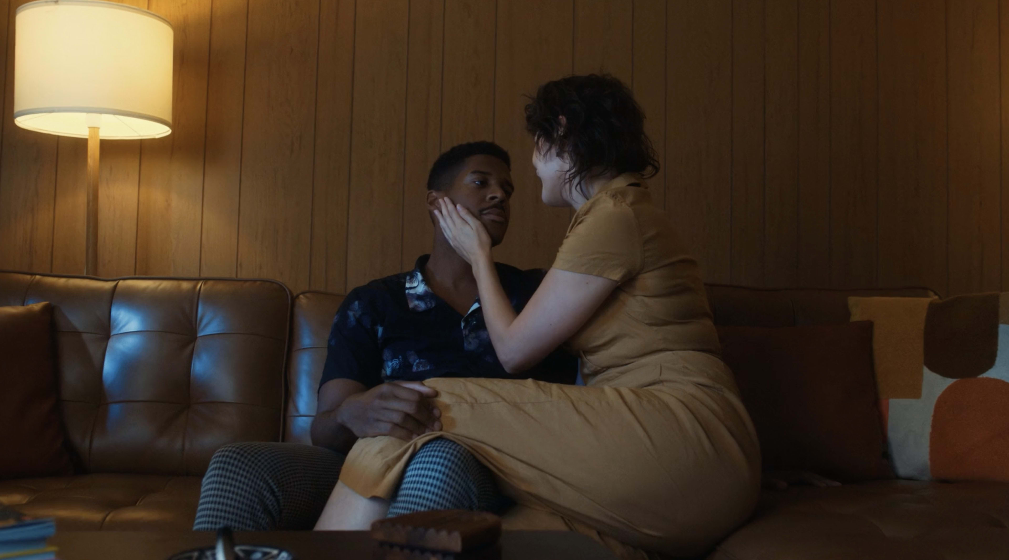 A man and a woman are seated closely on a couch in a warmly lit room, with the woman touching the man's face.