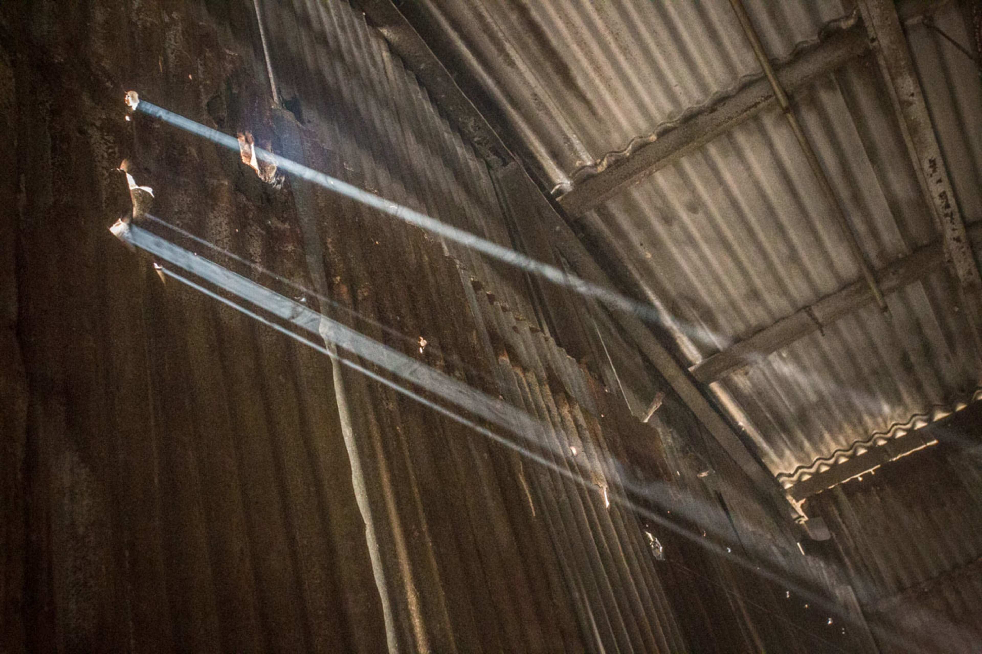 Sunlight filters through gaps in a corrugated metal roof, creating beams of light against the rusty walls of an interior space.