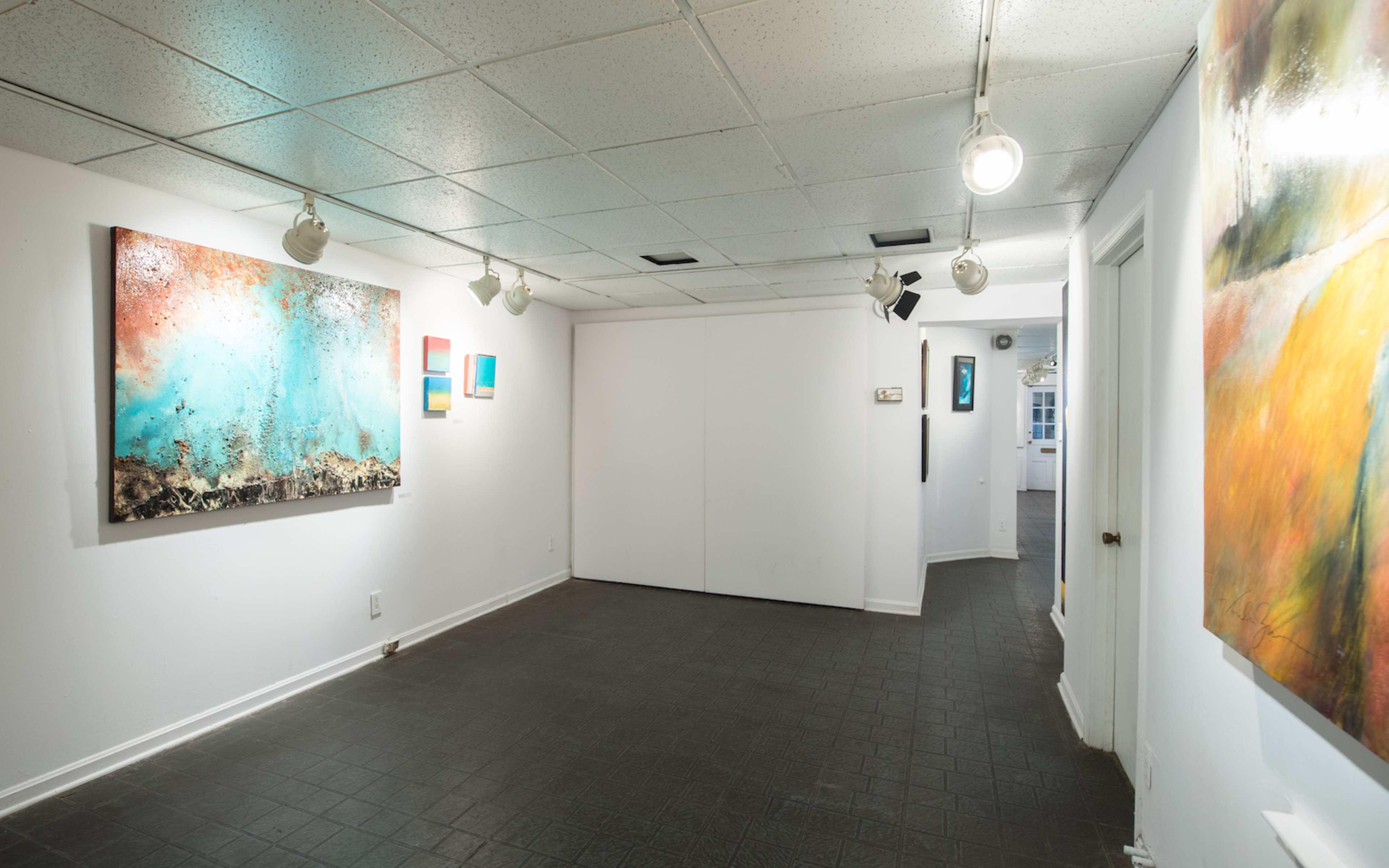 An empty art gallery with white walls, track lighting, and various colorful artworks displayed.