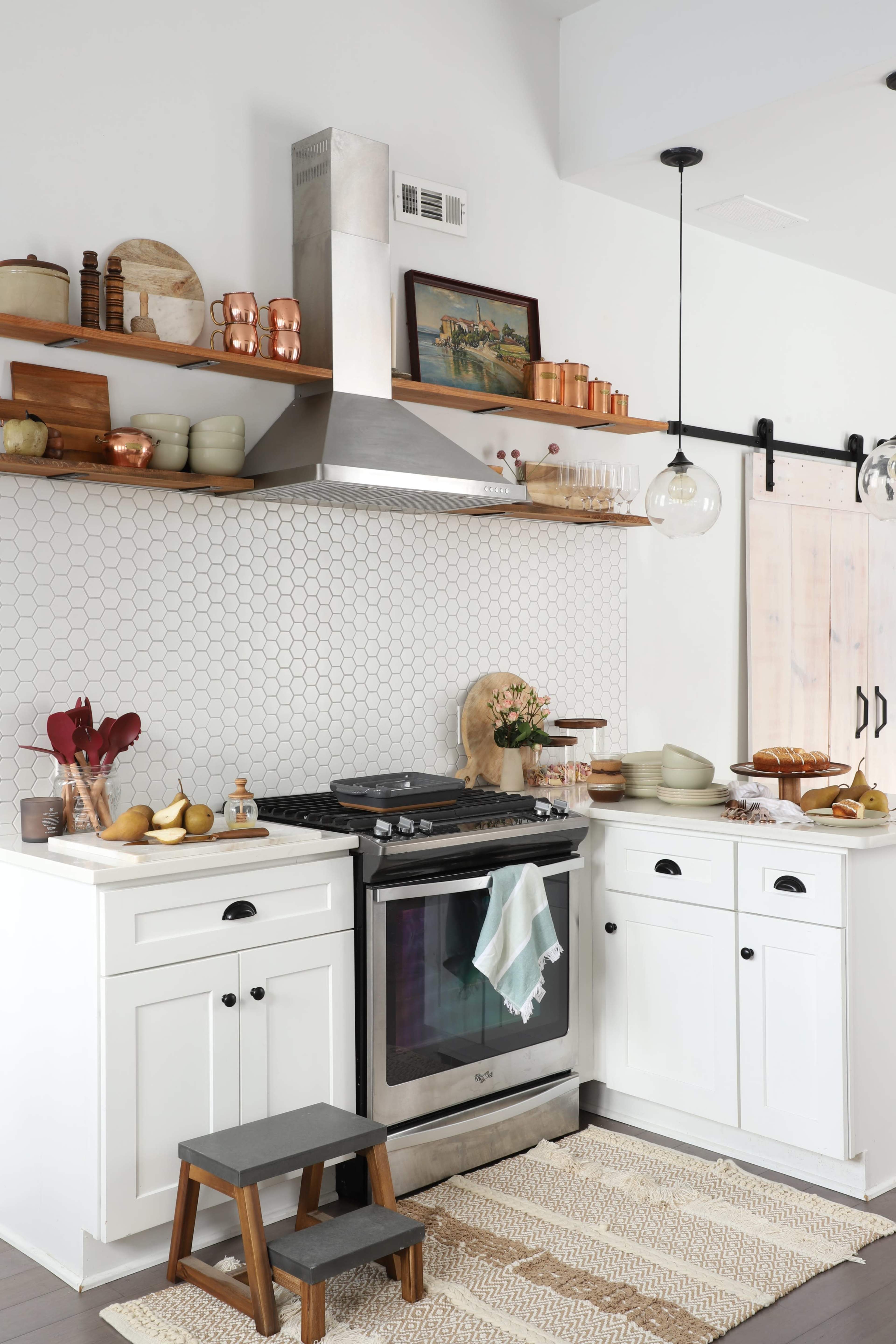 A modern kitchen featuring white cabinetry, a stainless steel stove, open shelving with decorative items, and a hexagonal tile backsplash.