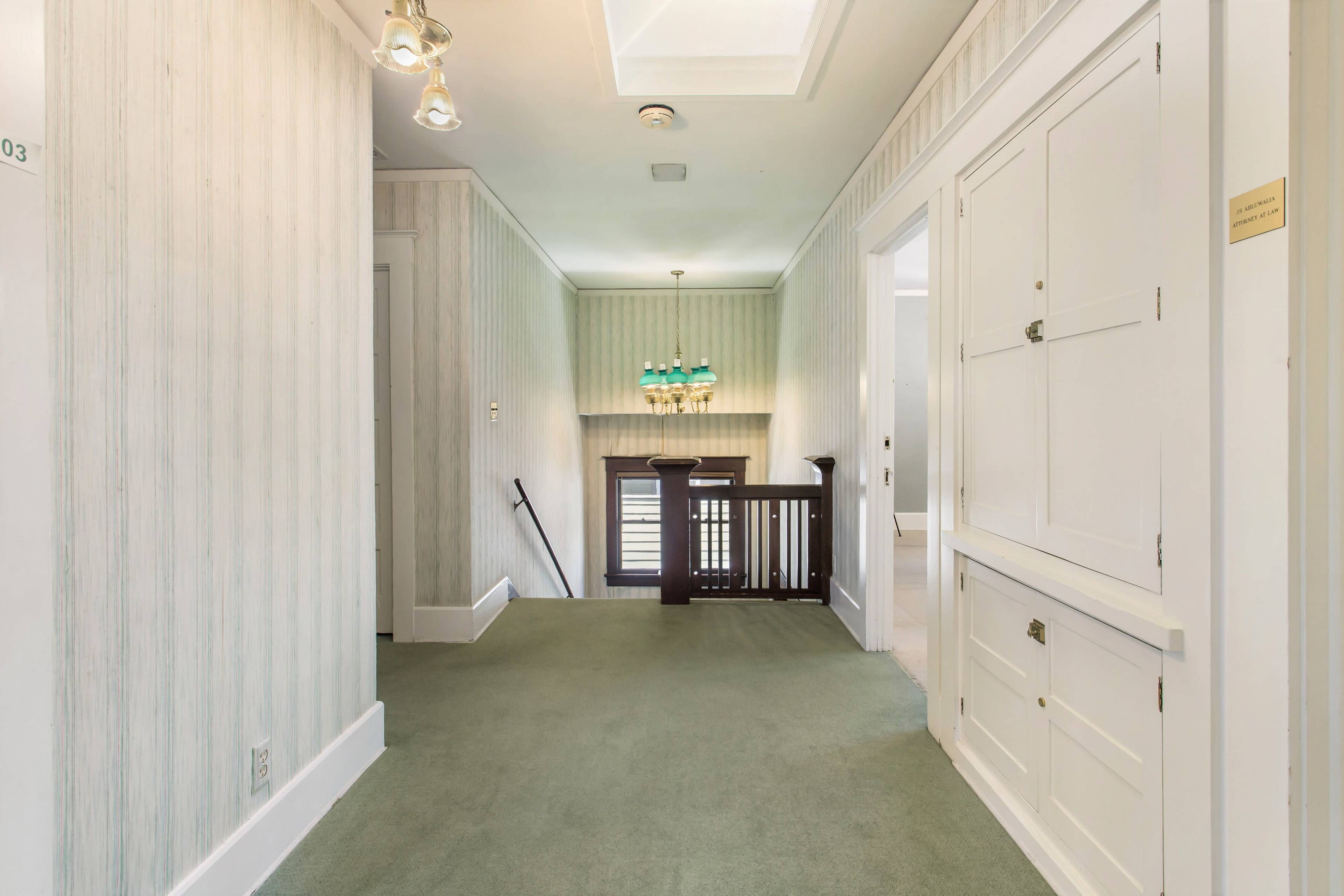 The image shows a hallway with green carpet, light-colored walls featuring striped wallpaper, and a wooden railing leading to a staircase.