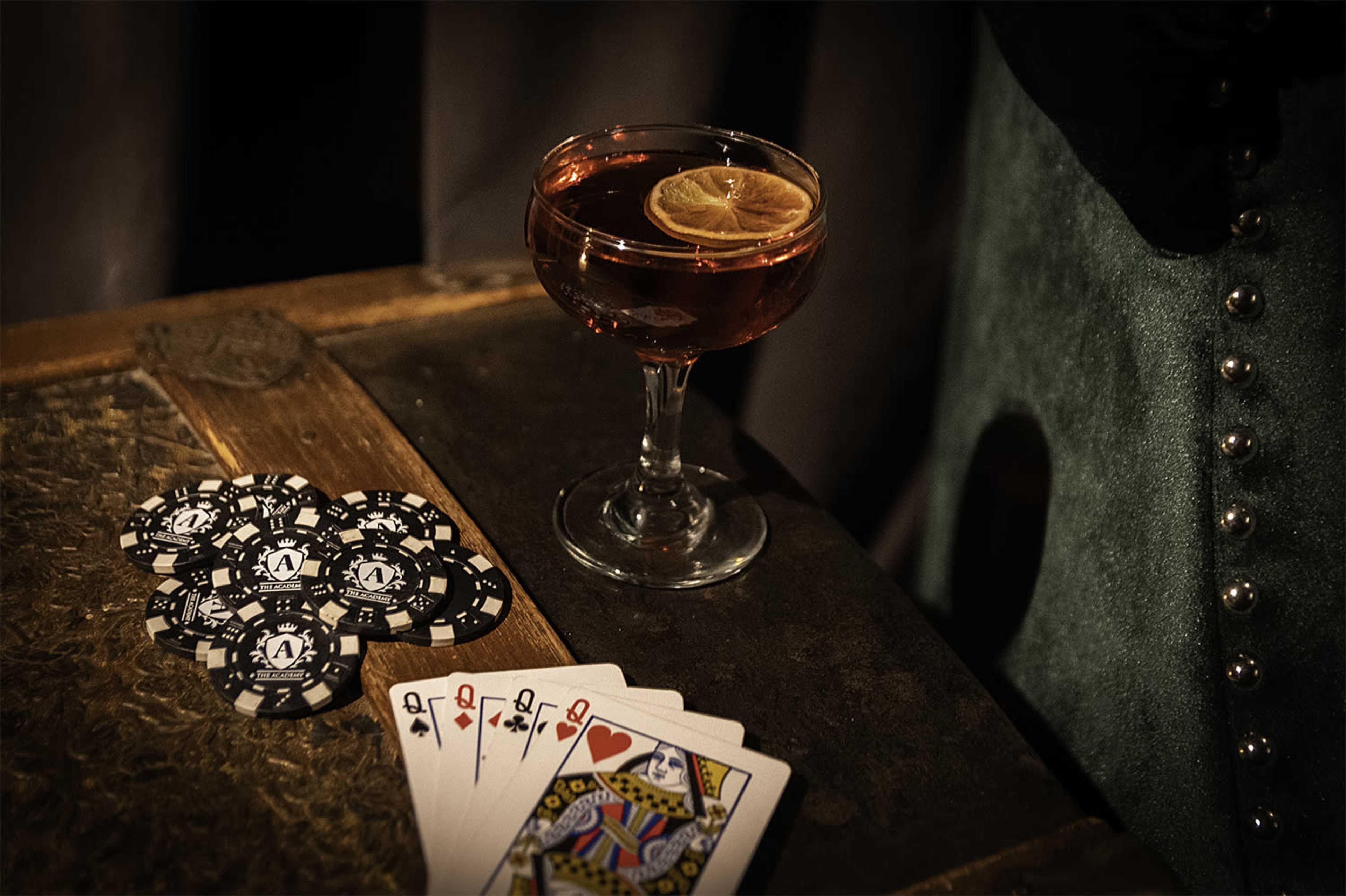 A cocktail glass with a slice of lemon sits next to a fan of playing cards and a stack of black poker chips on a wooden table.