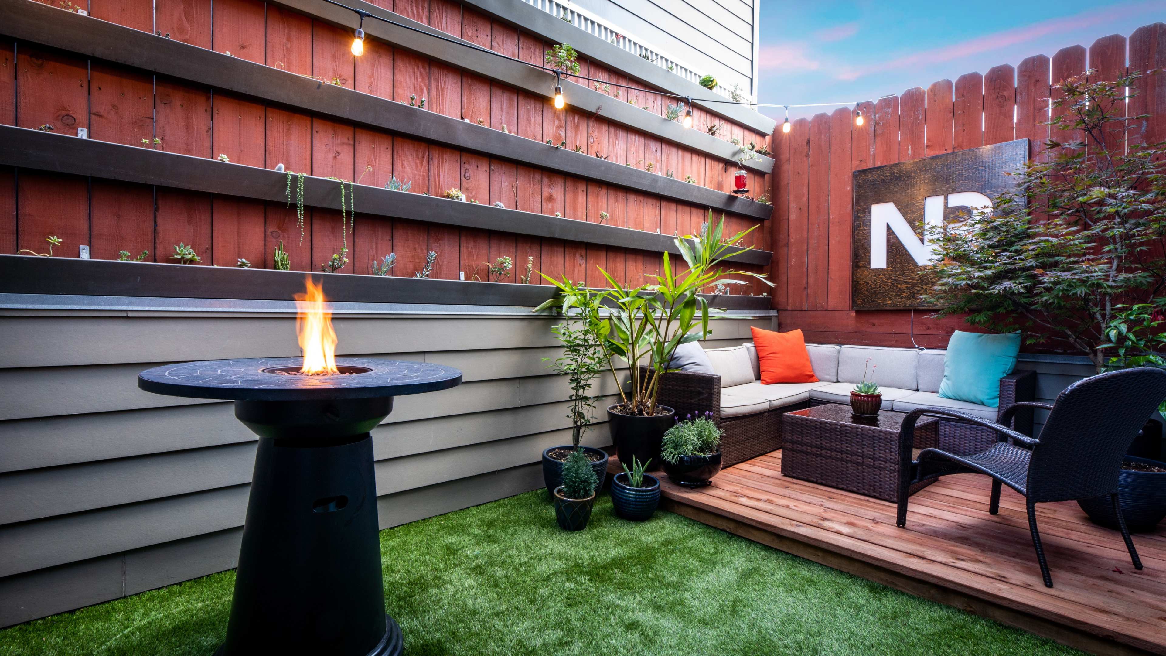 A small patio featuring a gas firepit, a seating area with colorful cushions, and various potted plants against a brown wooden fence.