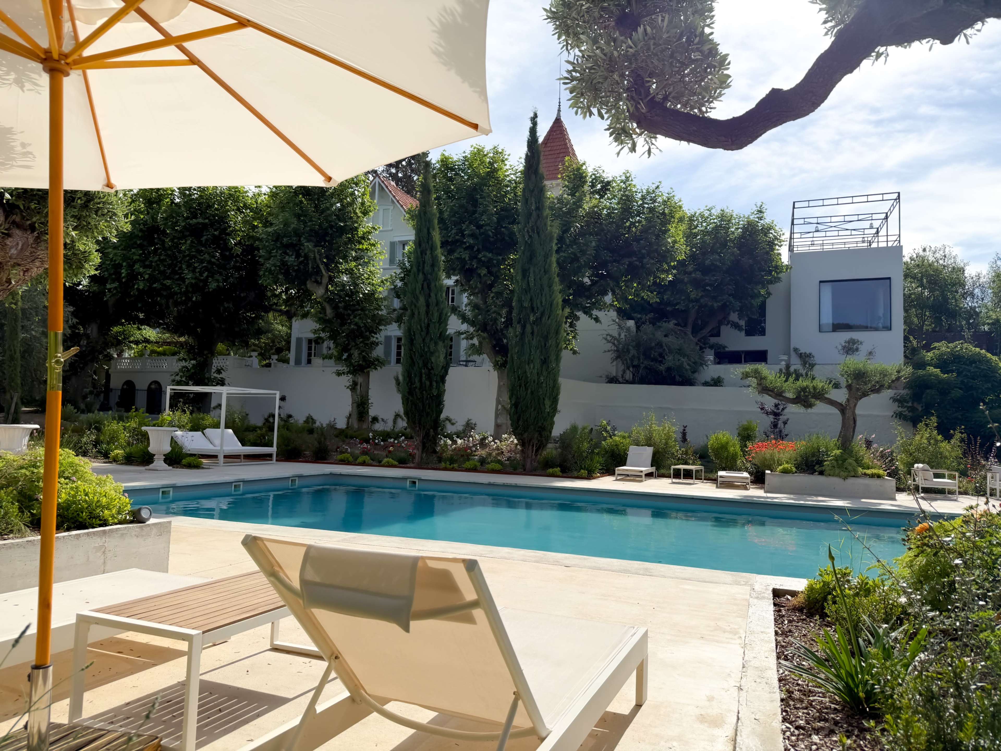 A swimming pool is surrounded by greenery and lounge chairs, with an umbrella providing shade.