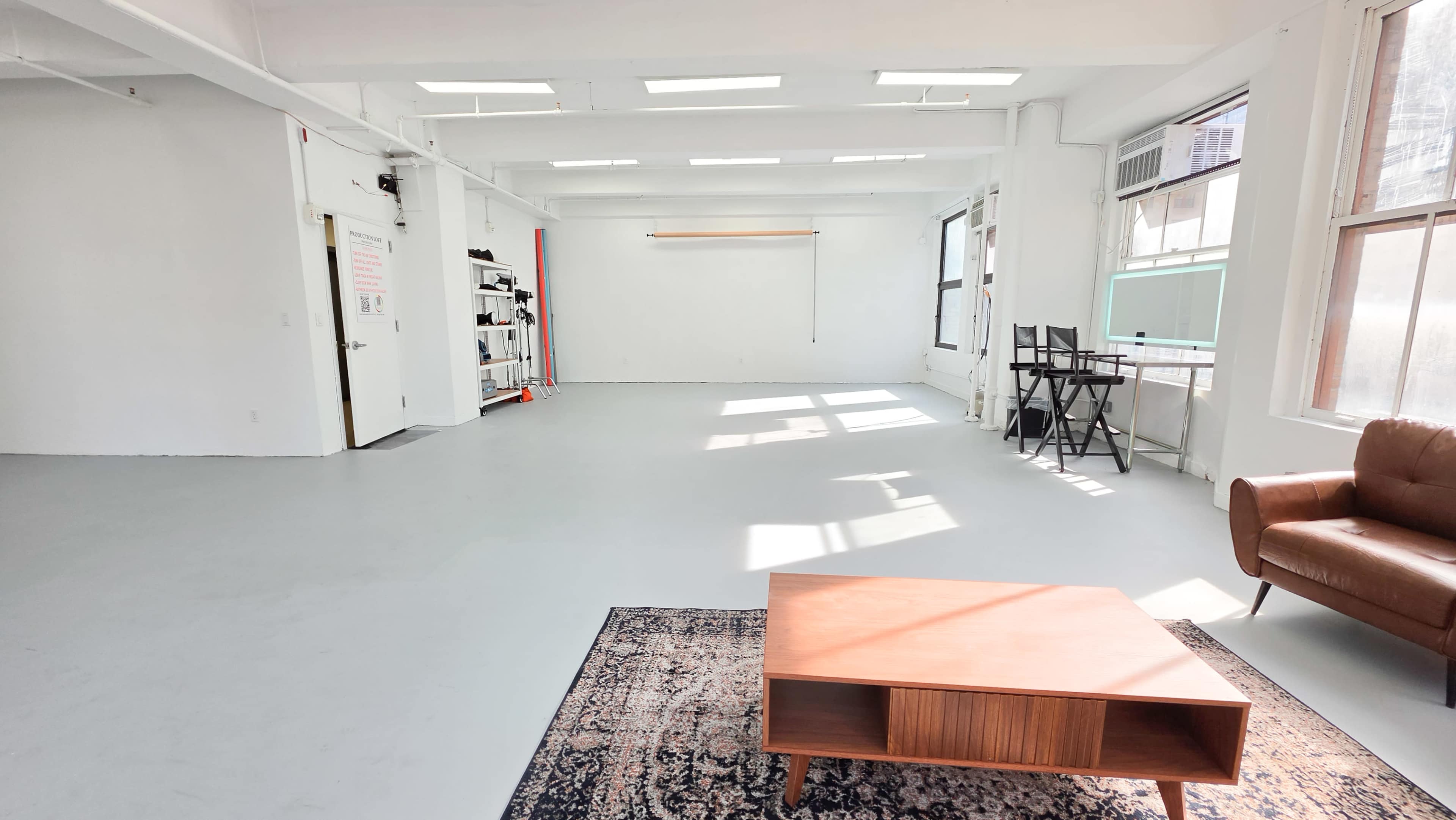 The image shows a spacious, well-lit room with white walls, a large window, a brown sofa, a wooden coffee table, and a few chairs near a desk.