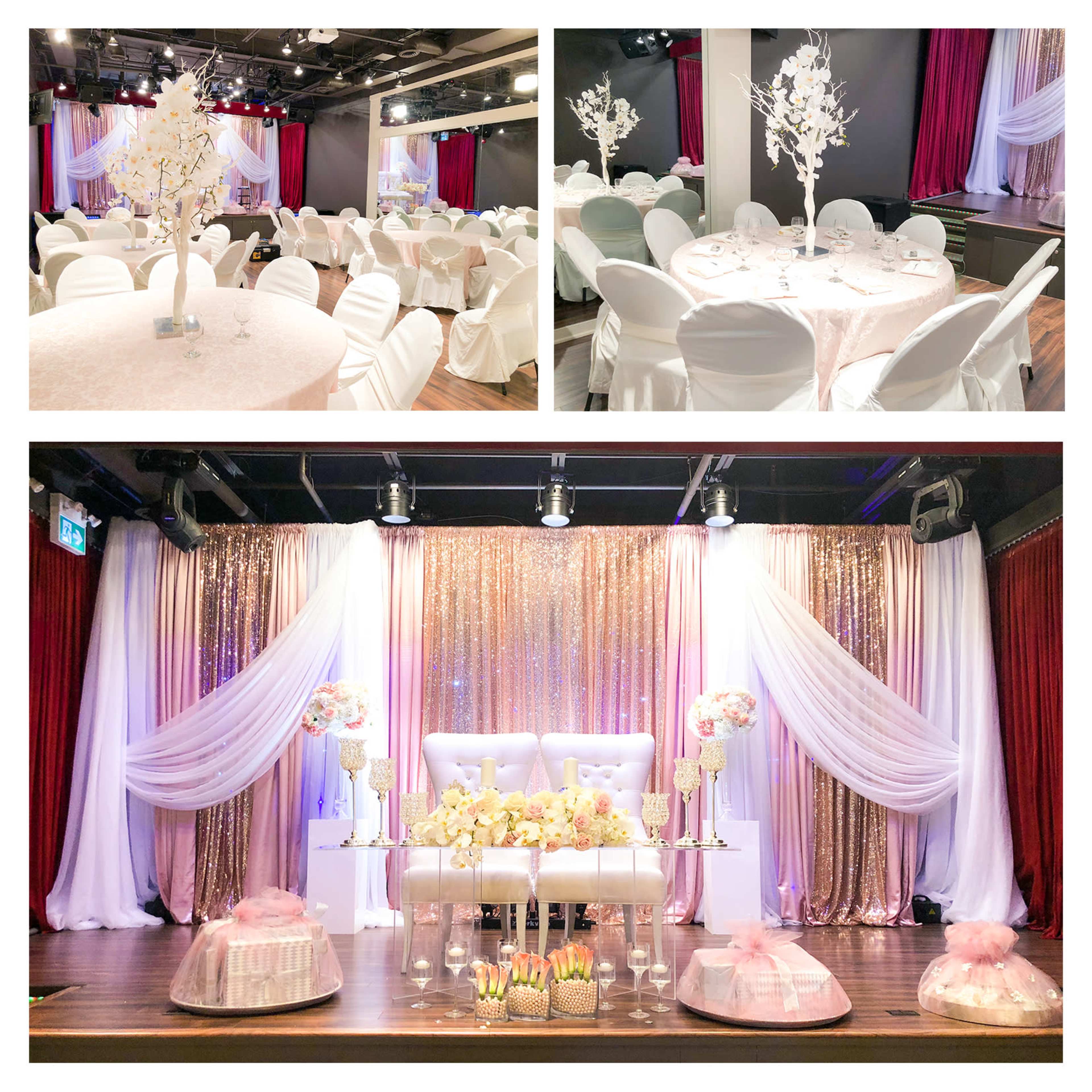 An elegantly decorated event space with round tables, white chairs, and a staged area featuring floral arrangements and draped fabrics.