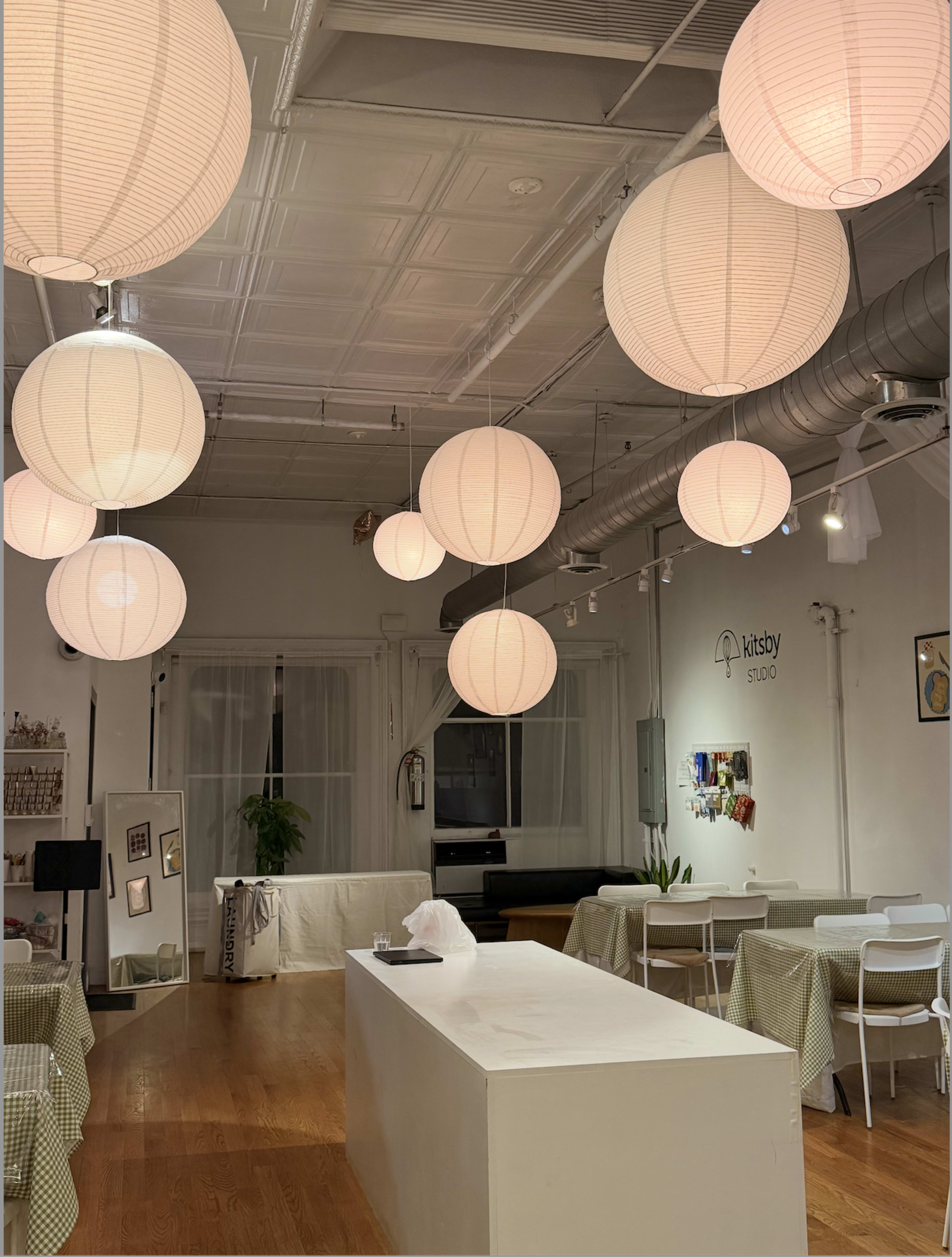 The image shows a well-lit studio space with round paper lanterns hanging from the ceiling, a central white table, and various seating arrangements.