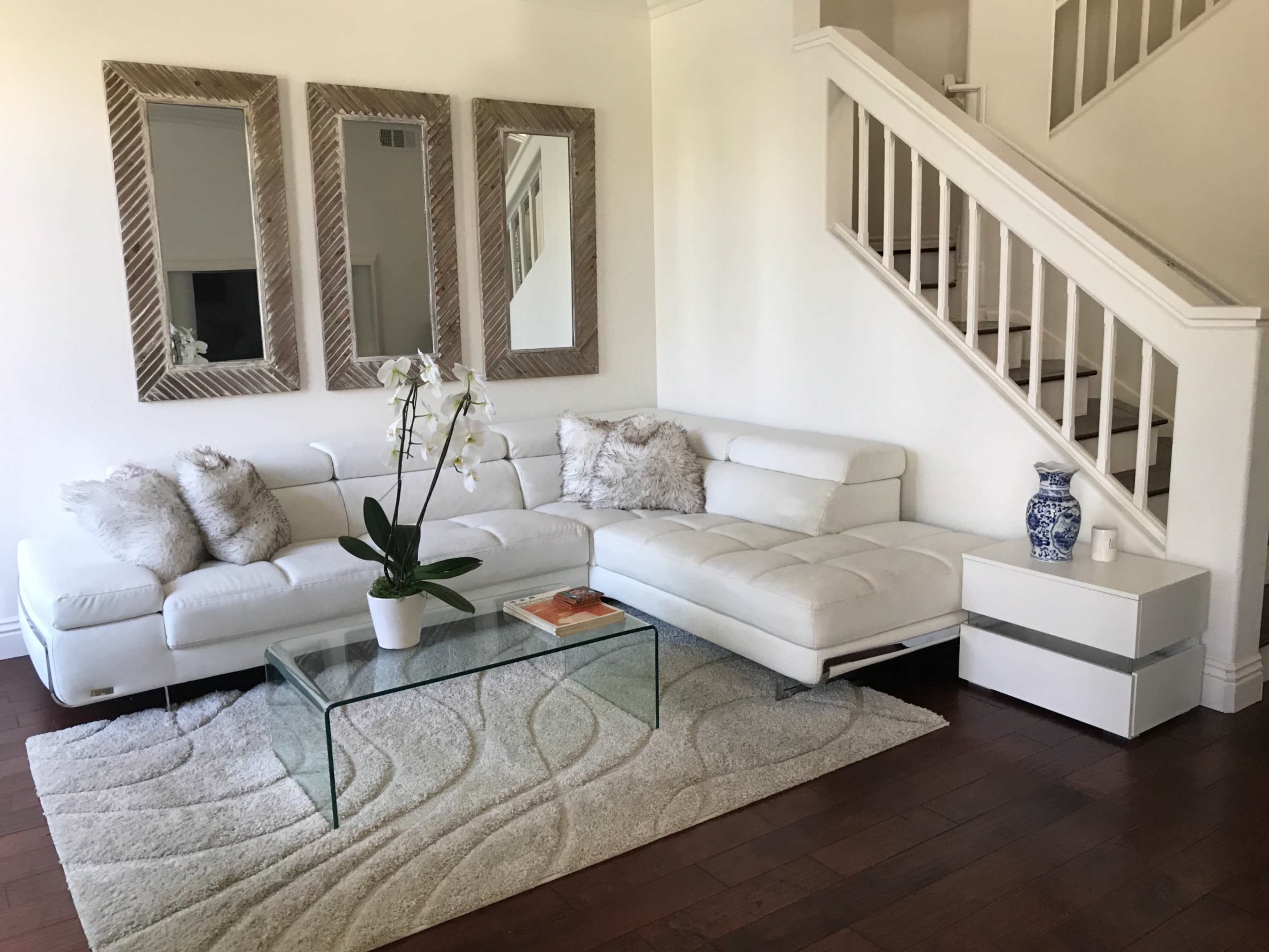 A modern living room with a white sectional sofa, a glass coffee table on a patterned rug, and three mirrors on the wall above.