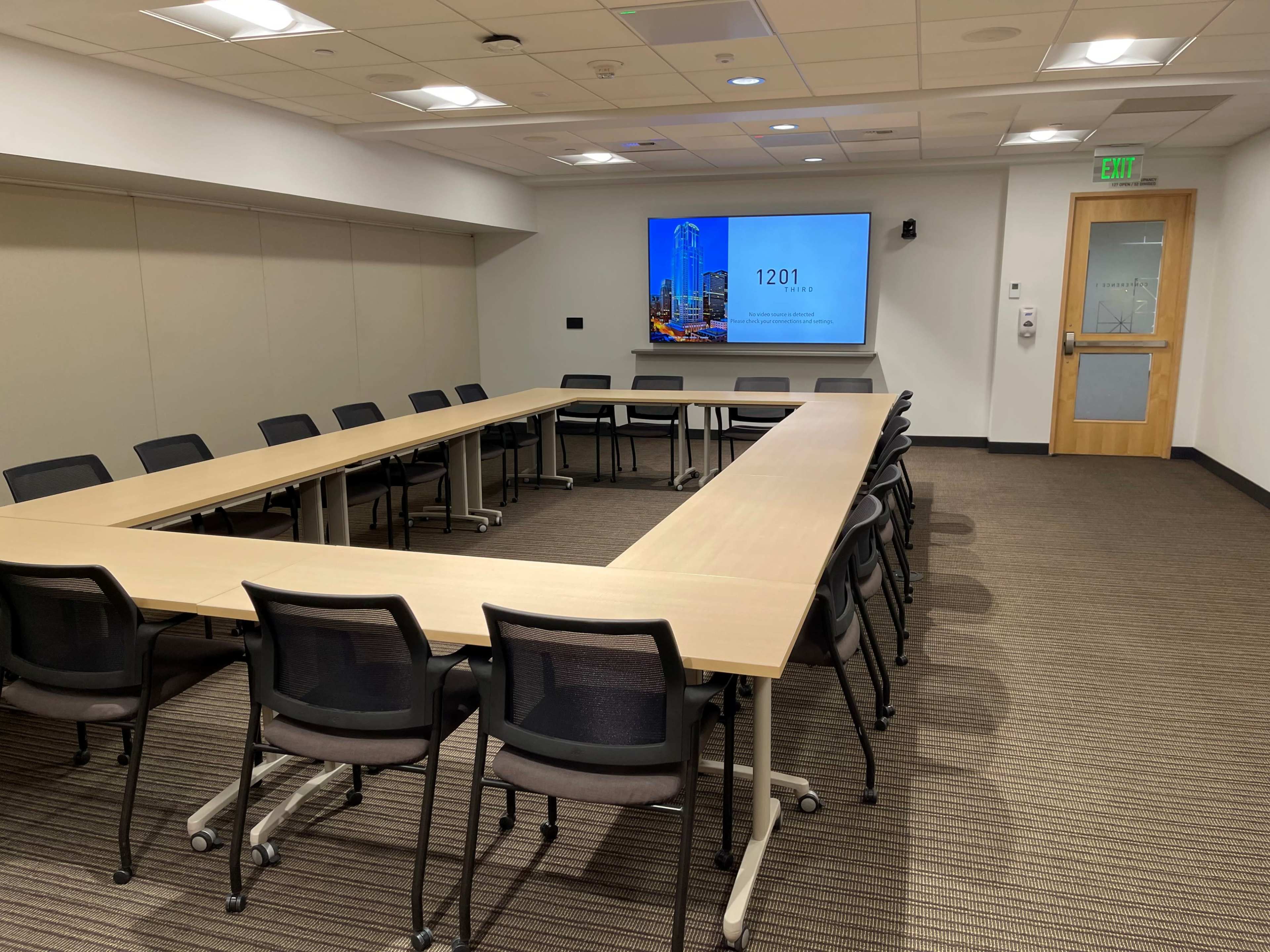 A large conference room features a rectangular table surrounded by chairs and a wall-mounted screen displaying the number "1201."