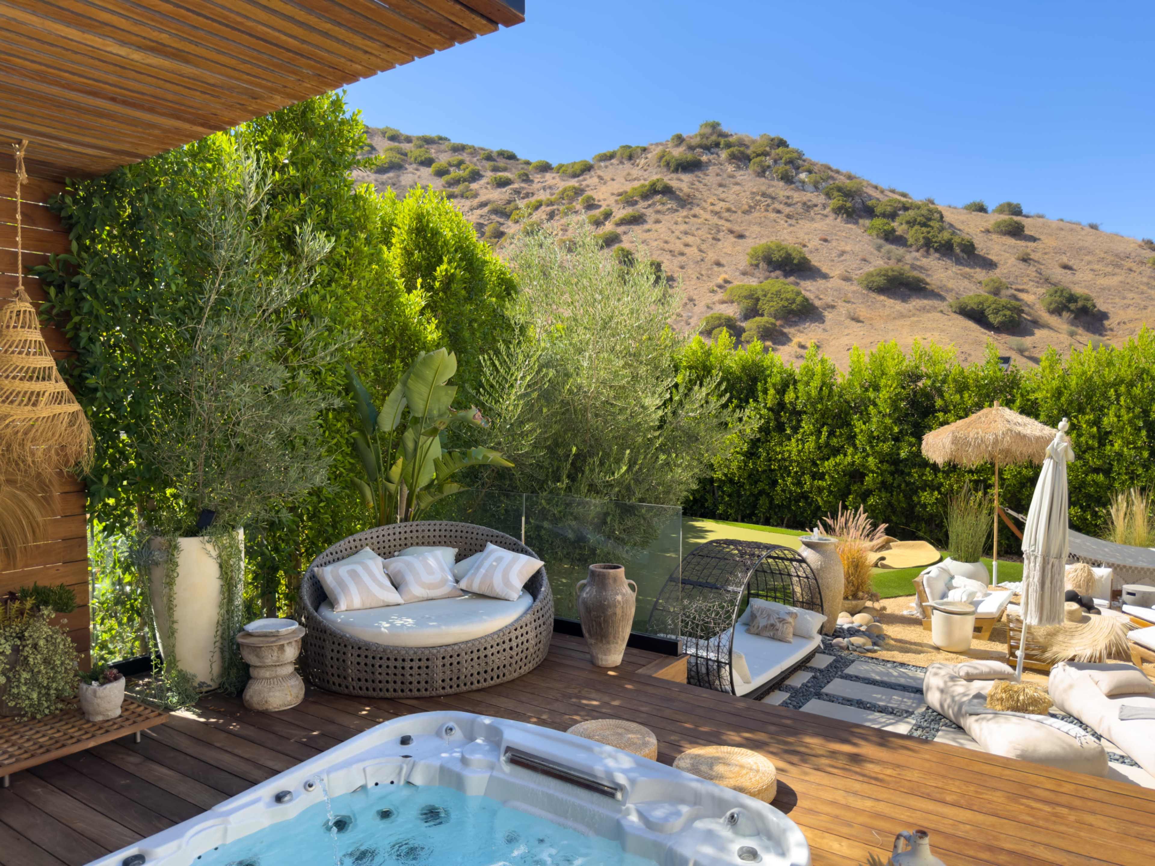 A modern outdoor space features a hot tub, comfortable seating, and lush greenery against a hillside backdrop.