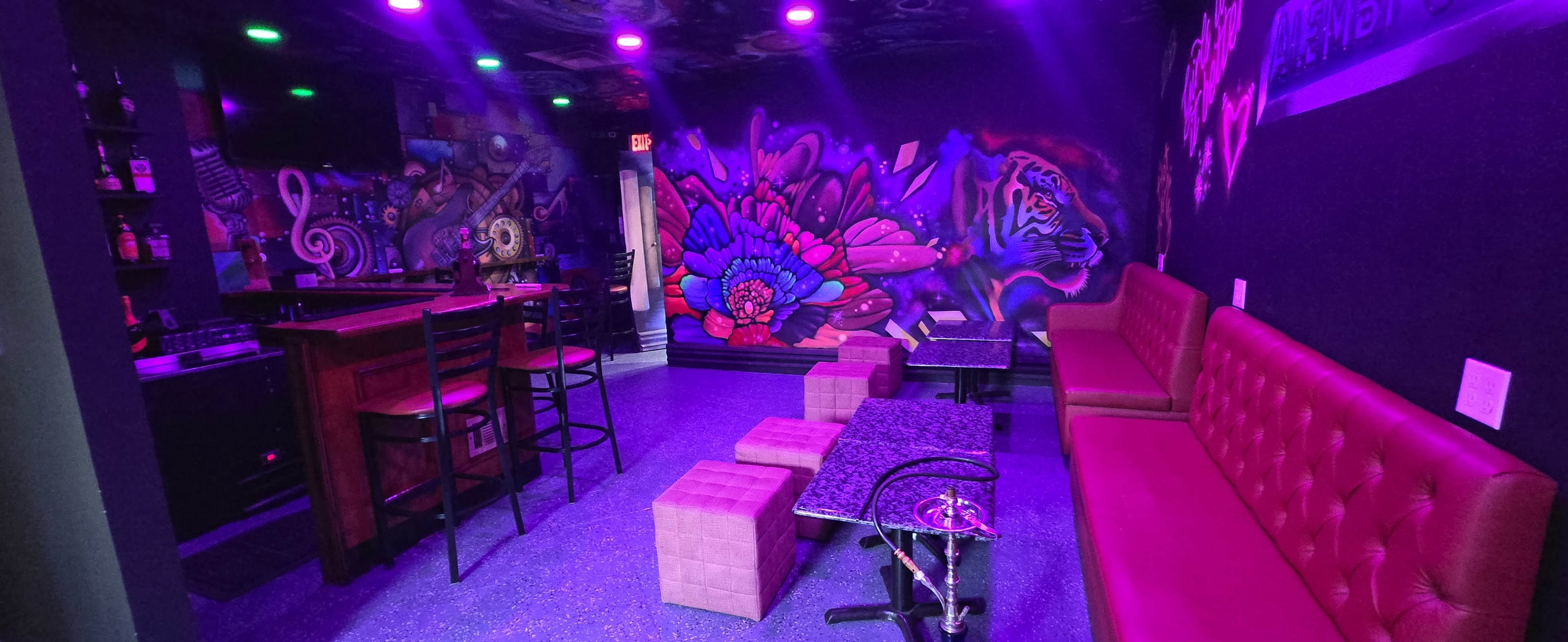The image shows a cozy lounge with vibrant murals on the walls, purple lighting, and a bar area with seating.