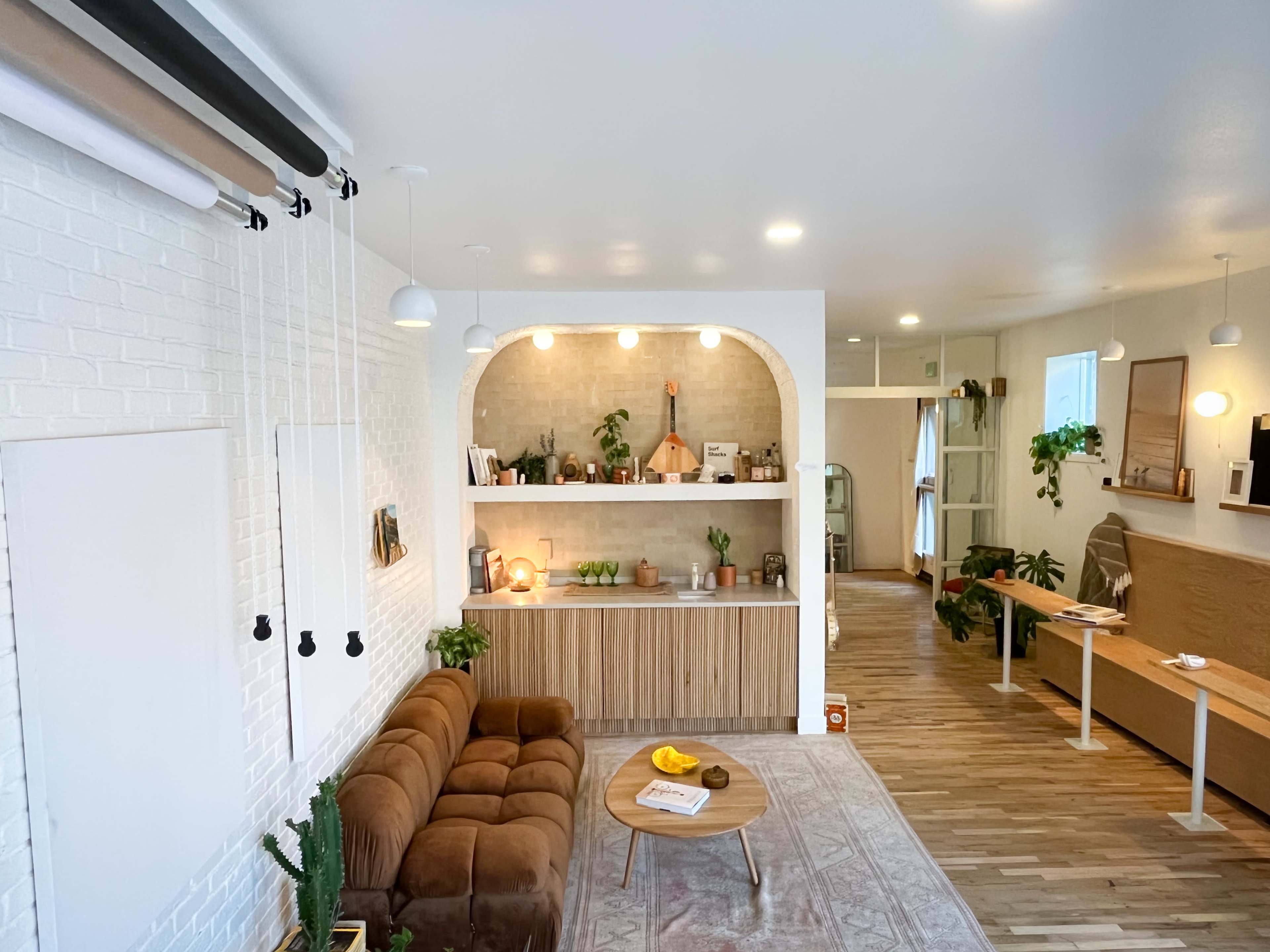 A modern, open-concept living space with a brown sofa, a central wooden table, and a kitchen area featuring plants and various decorative items.