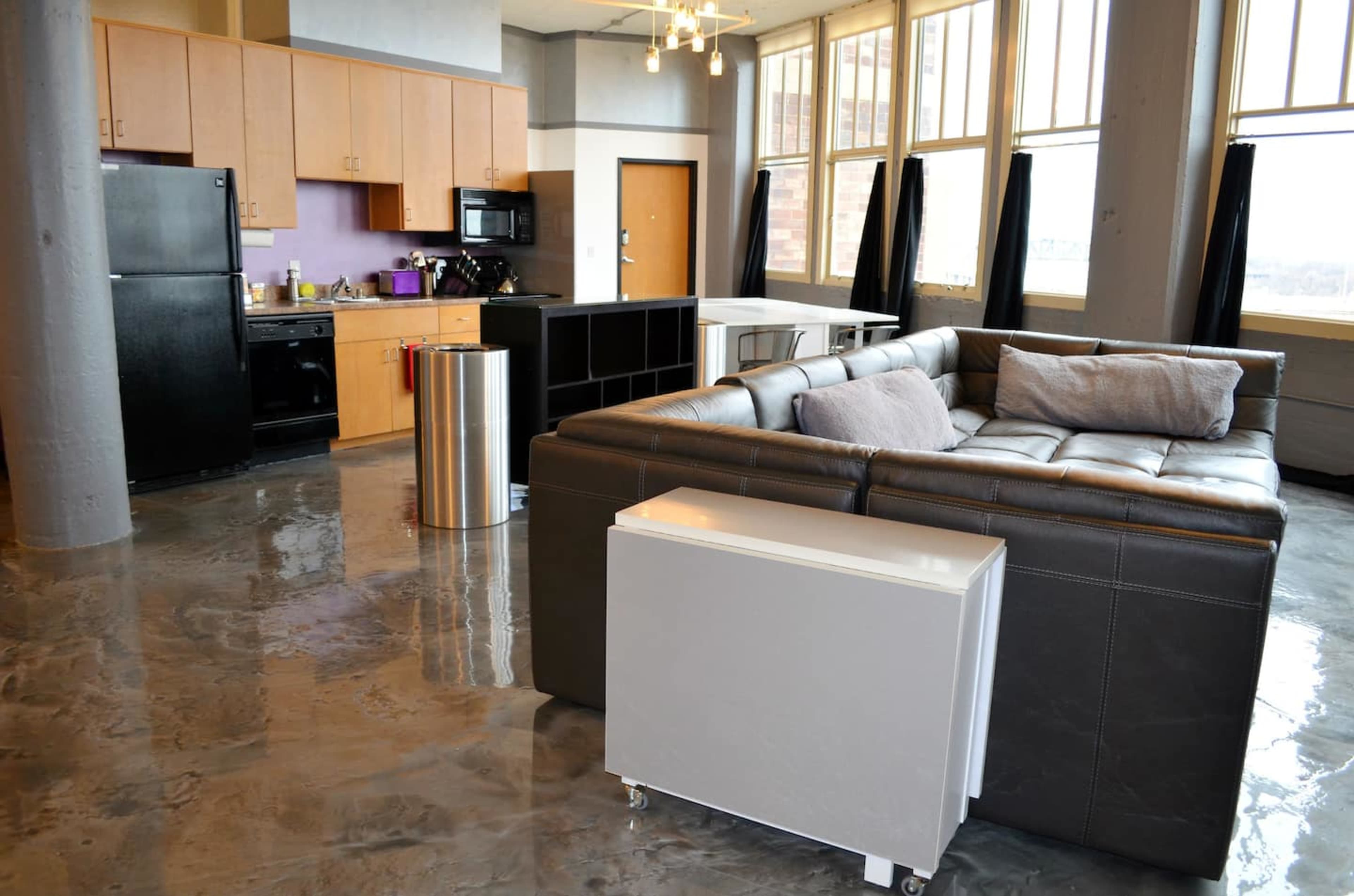 A modern kitchen and living area featuring a dark leather sofa, stainless steel appliances, and polished concrete floors.