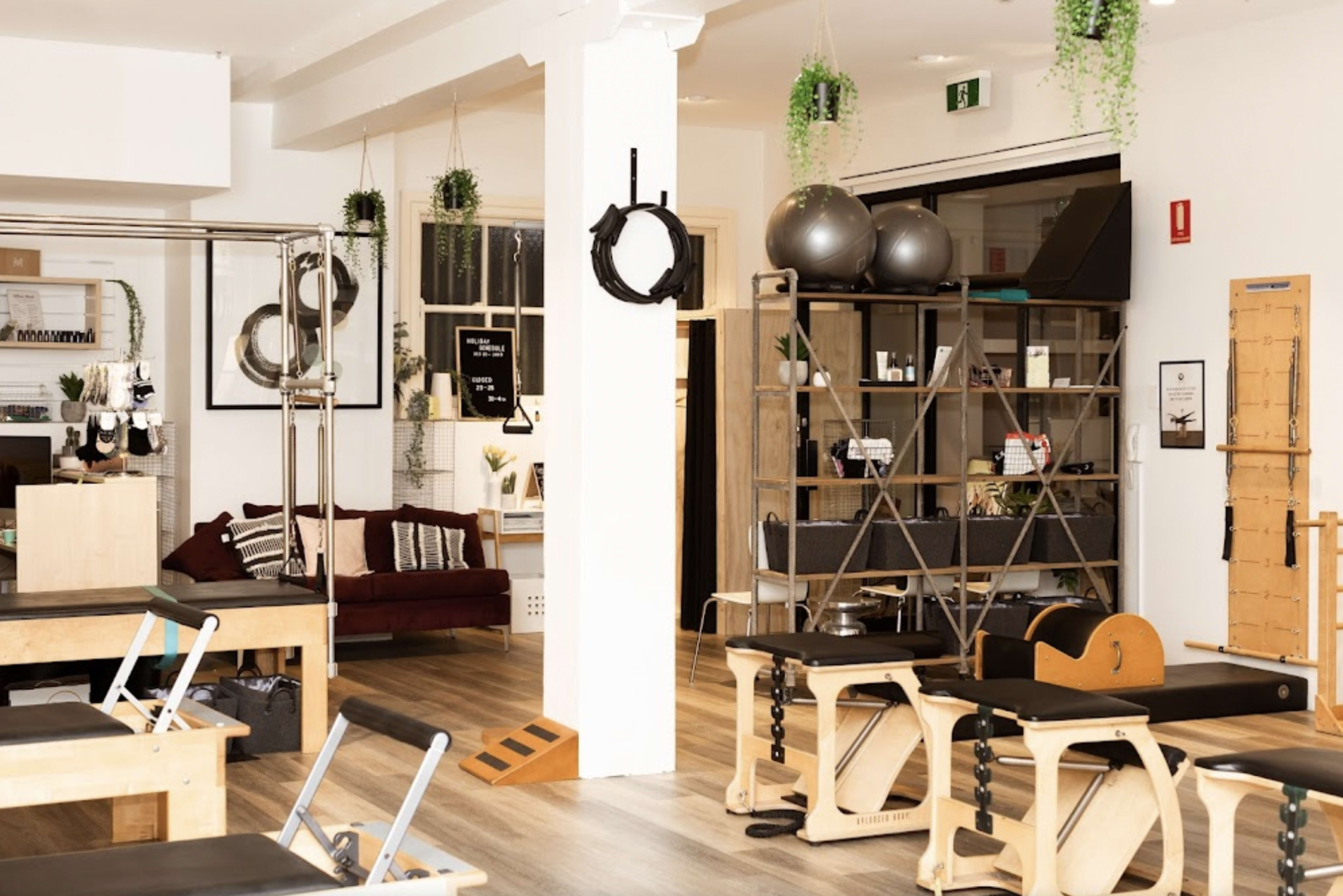 A spacious Pilates studio with various exercise equipment, a seating area with a couch, and greenery hanging from the ceiling.