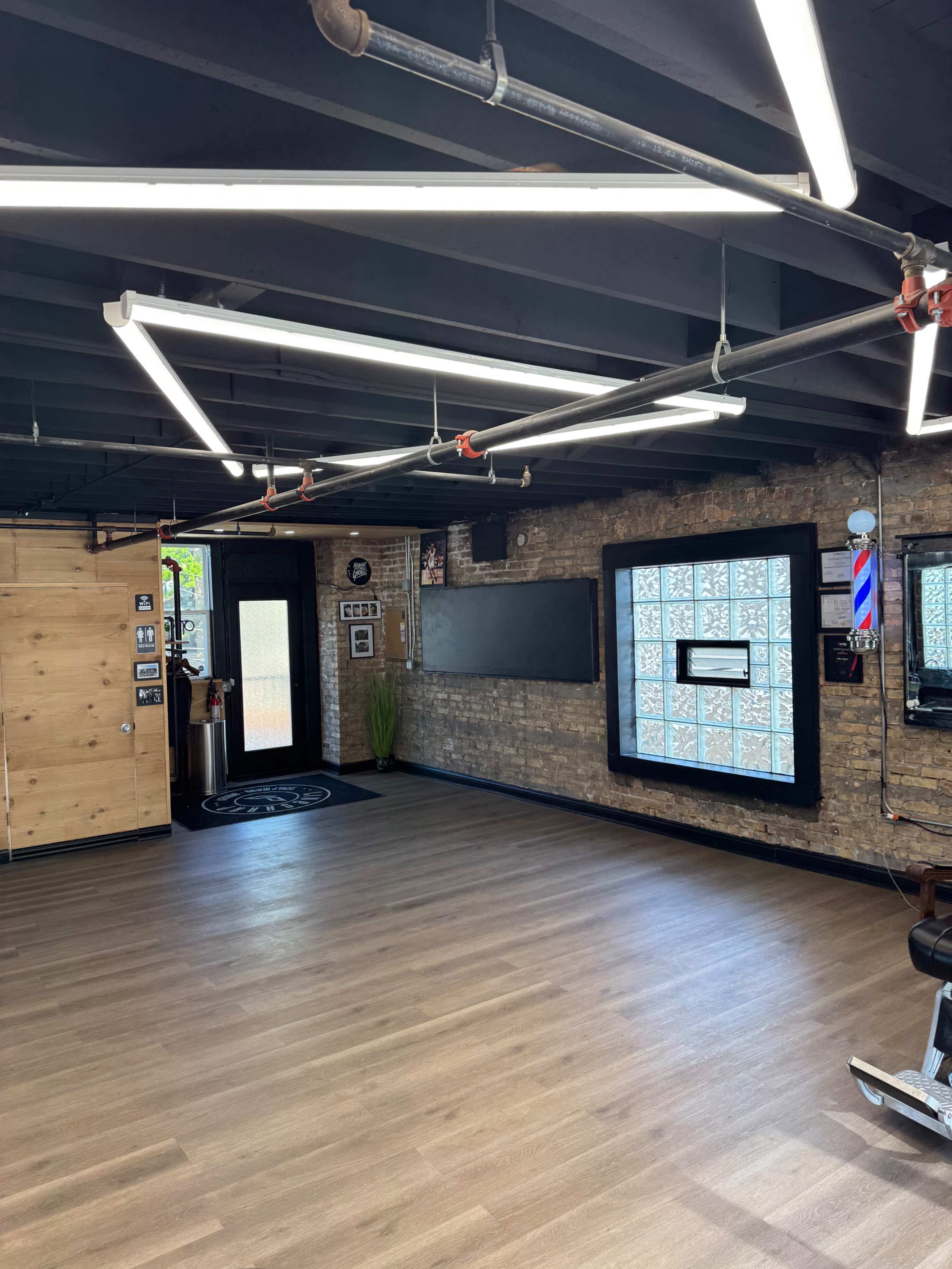 The image shows the interior of a barbershop featuring wooden walls, modern lighting, and a sleek floor with a waiting area and a barber station.
