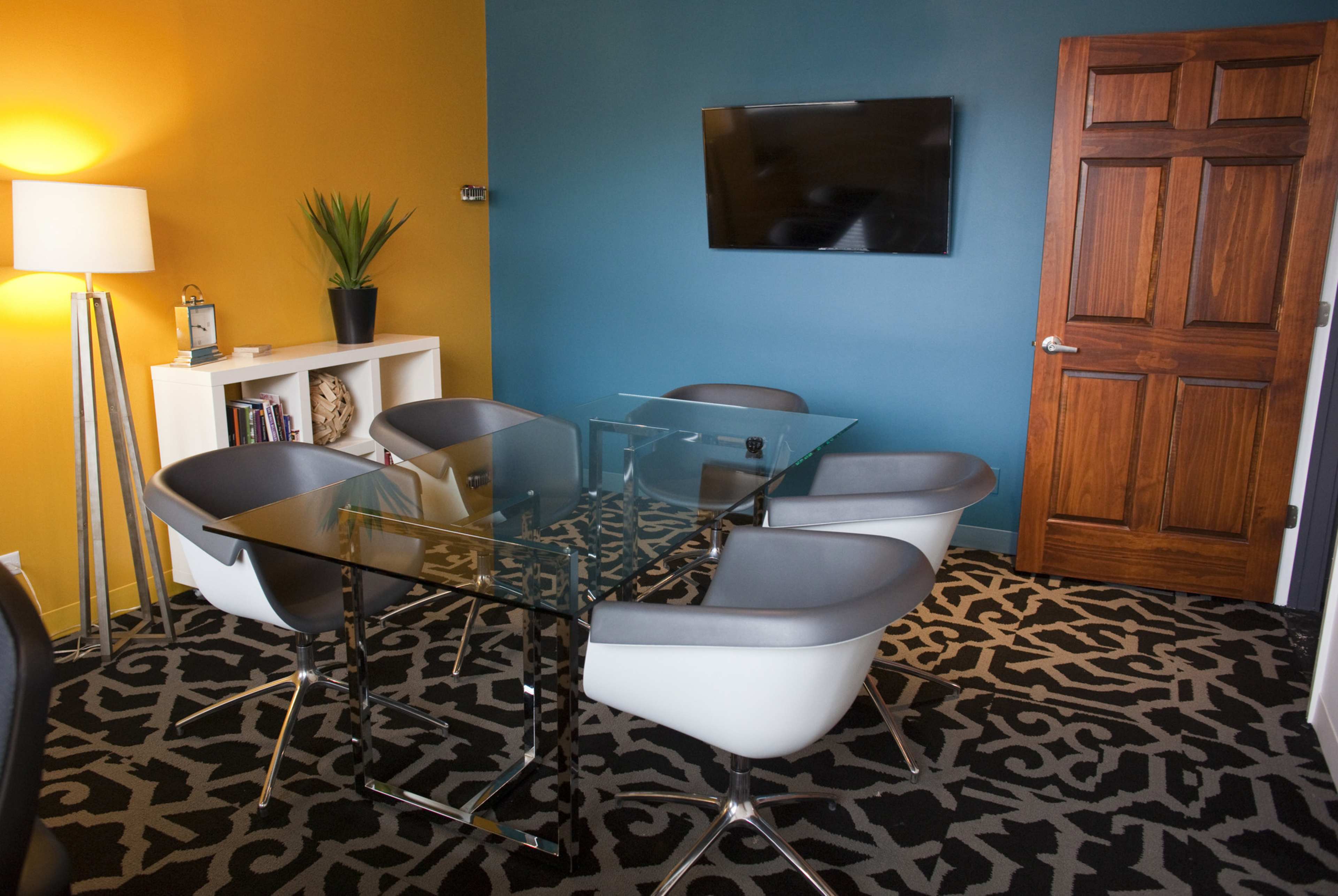 A modern conference room features a glass table surrounded by stylish chairs, with a television mounted on a wall and a lamp beside a plant in one corner.
